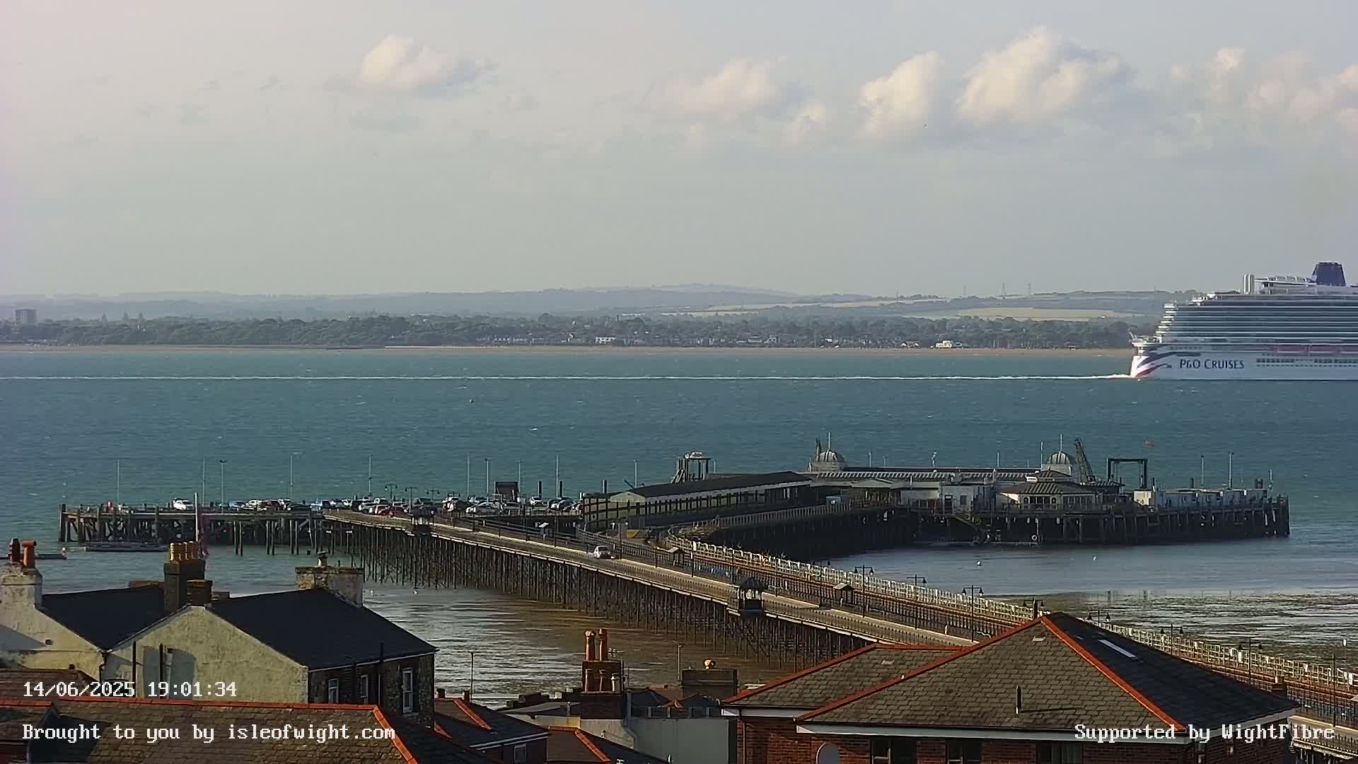 A long pier extends into a calm body of water, with a large cruise ship in the distance under a partly cloudy sky.