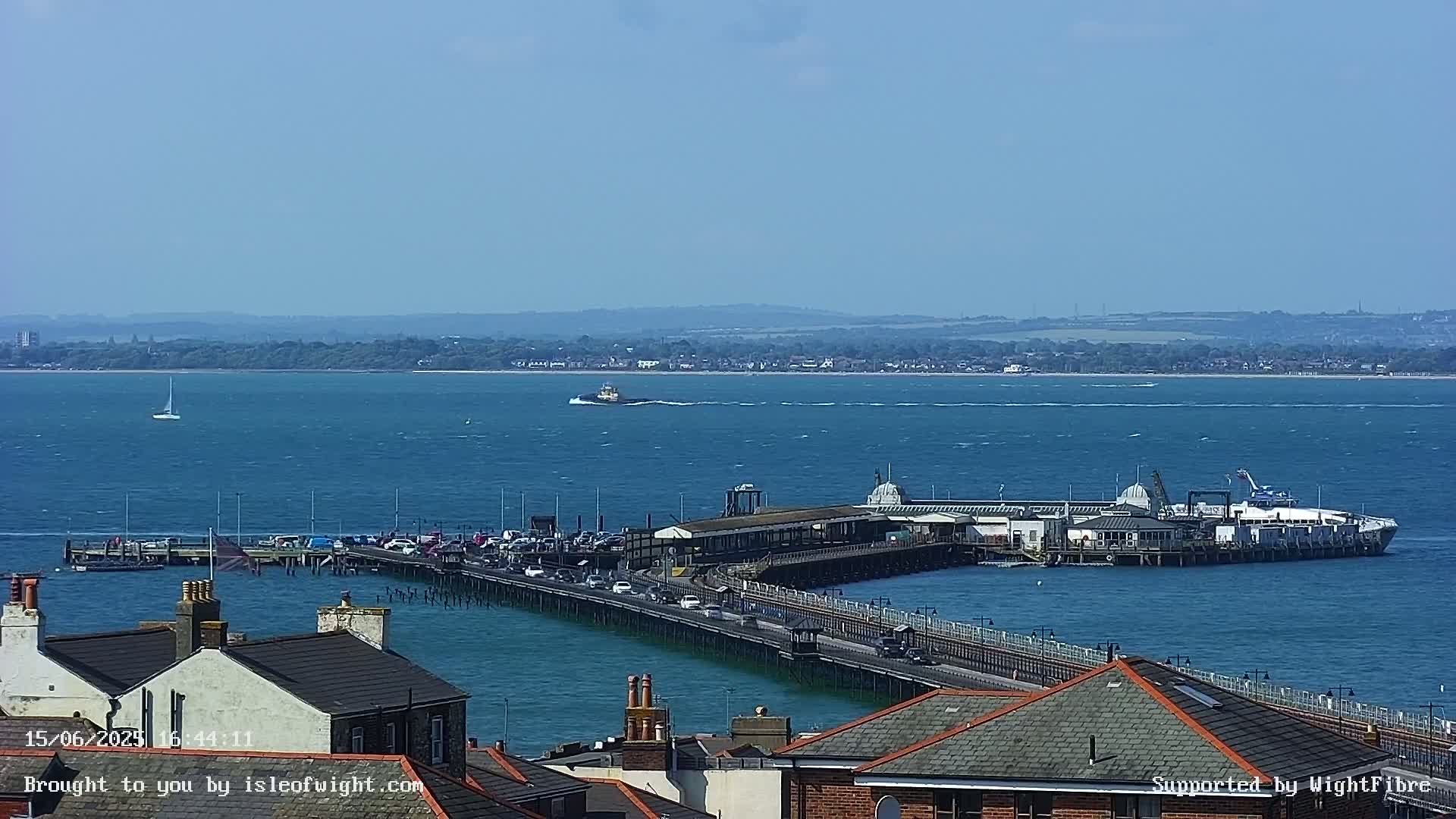 A long pier extends into a calm, blue body of water on a sunny day, with a town visible in the foreground and a distant shoreline in the background.