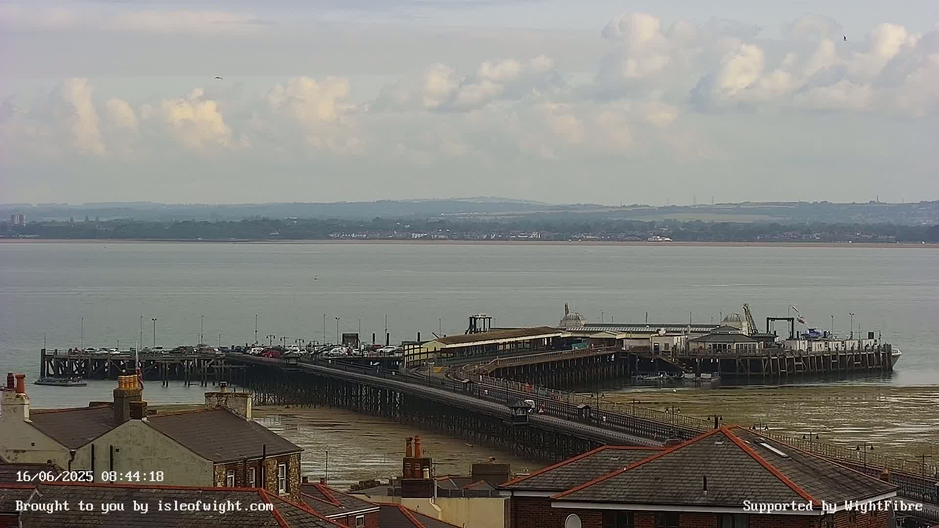 A long pier extends into a calm body of water under a partly cloudy sky, with buildings and rooftops visible in the foreground.