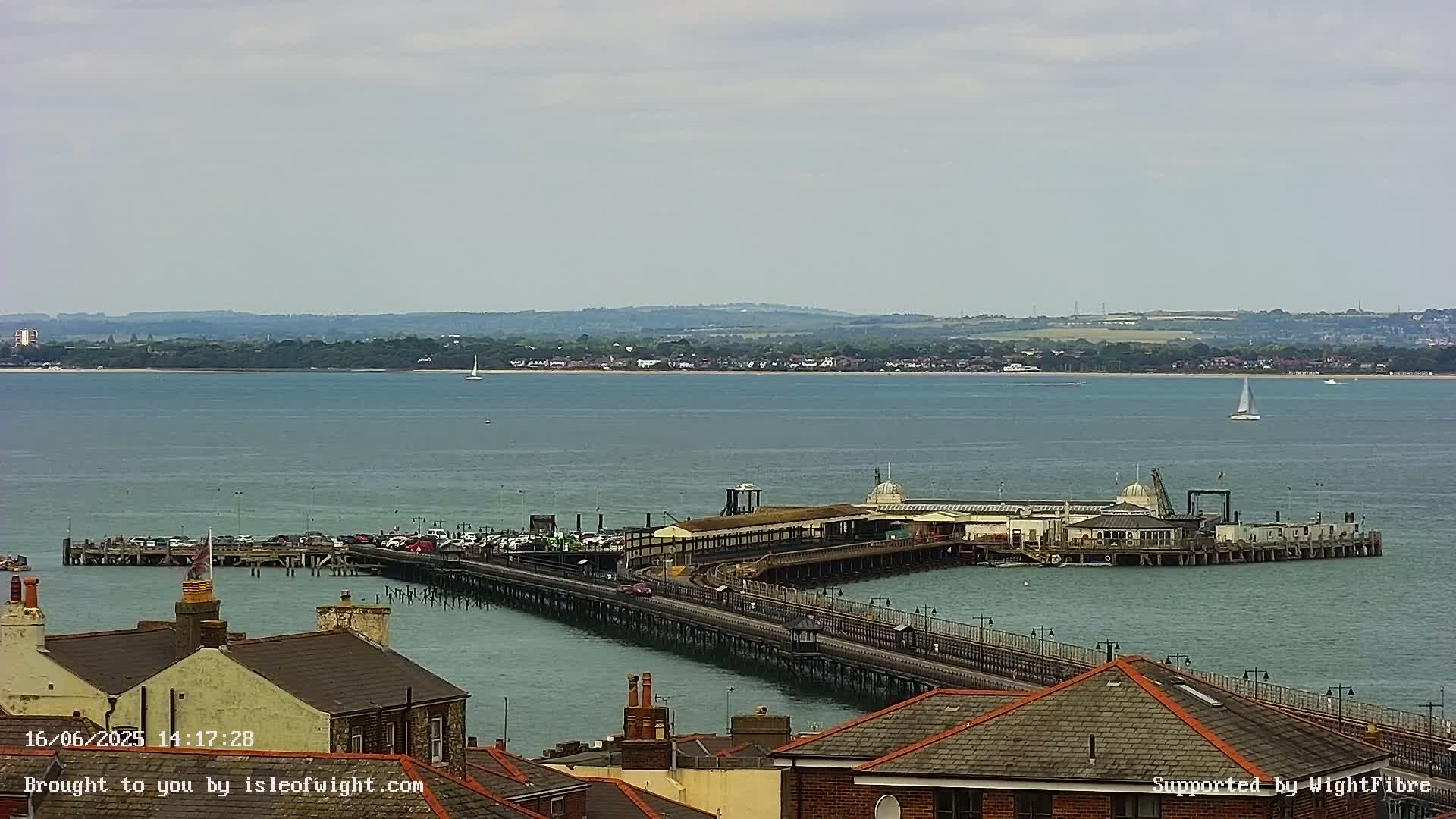 A long pier extends into a calm body of water under an overcast sky, with buildings and rooftops visible in the foreground and a distant shoreline in the background.