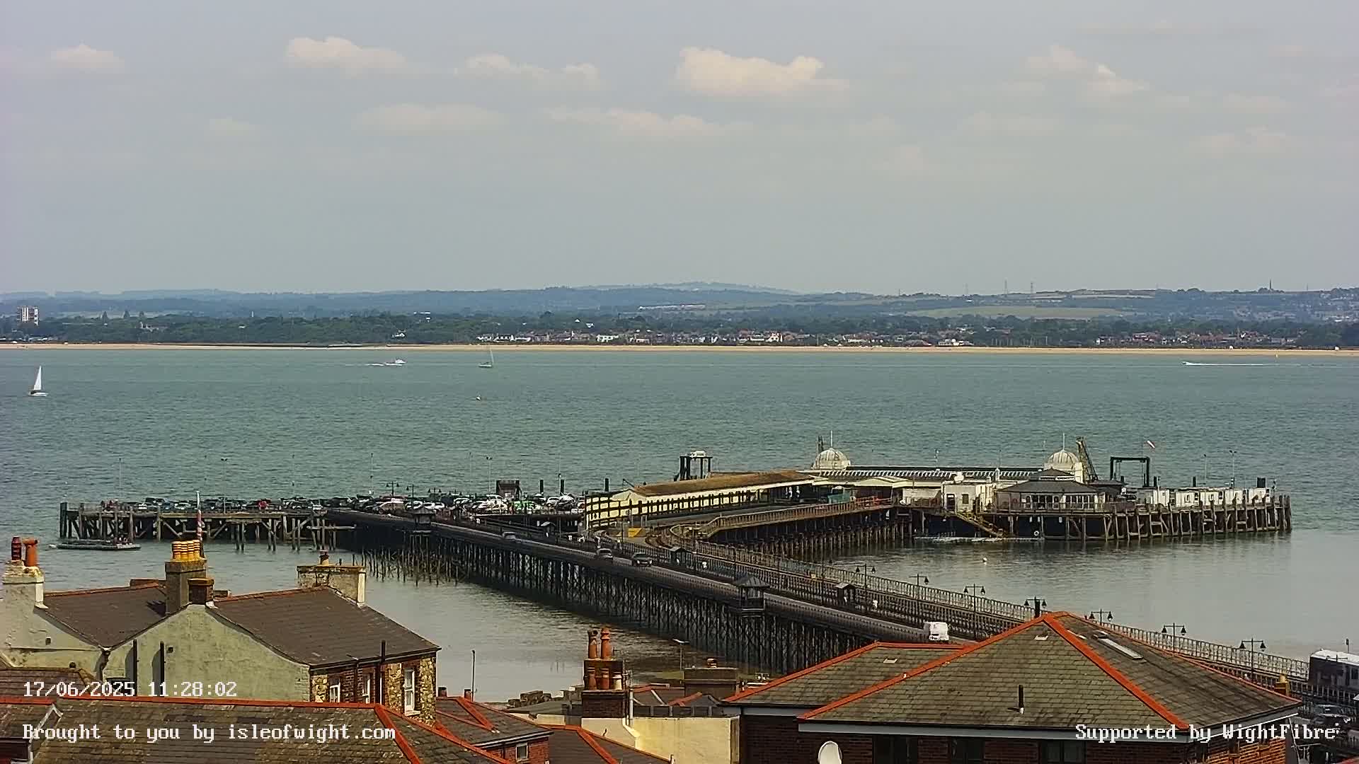 A long pier extends from a town into a calm body of water under a mostly cloudy sky, with a distant shoreline visible.