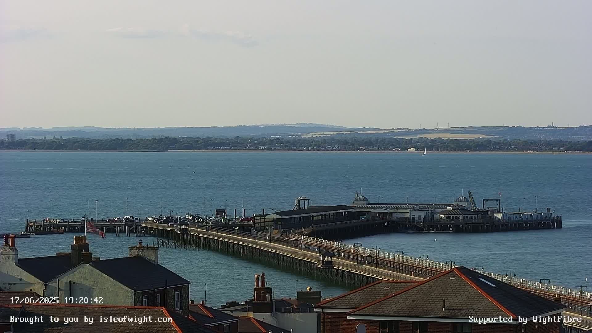 A long pier extends into a calm body of water, with a town and distant land visible under a clear sky.