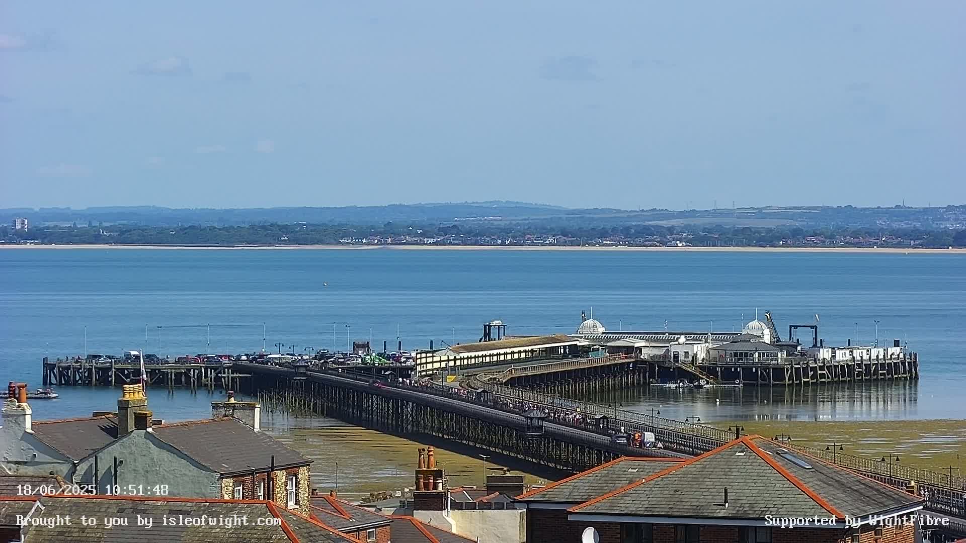 A long pier extends into a calm body of water under a clear sky, with buildings and a shoreline visible in the foreground and a distant landmass in the background.