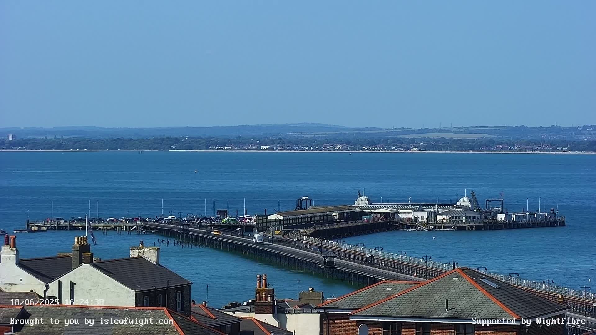 A long pier extends into a calm, blue body of water on a sunny day, with rooftops of buildings visible in the foreground.