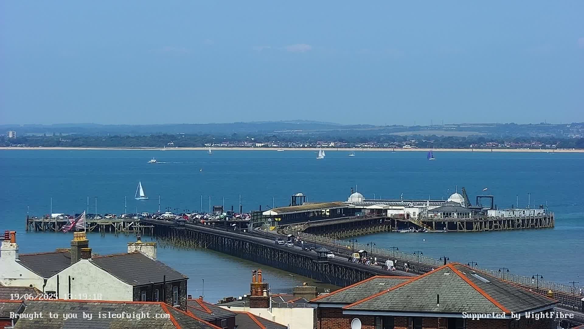 A long pier extends from a town into a calm, blue body of water on a sunny day, with several sailboats visible in the distance.