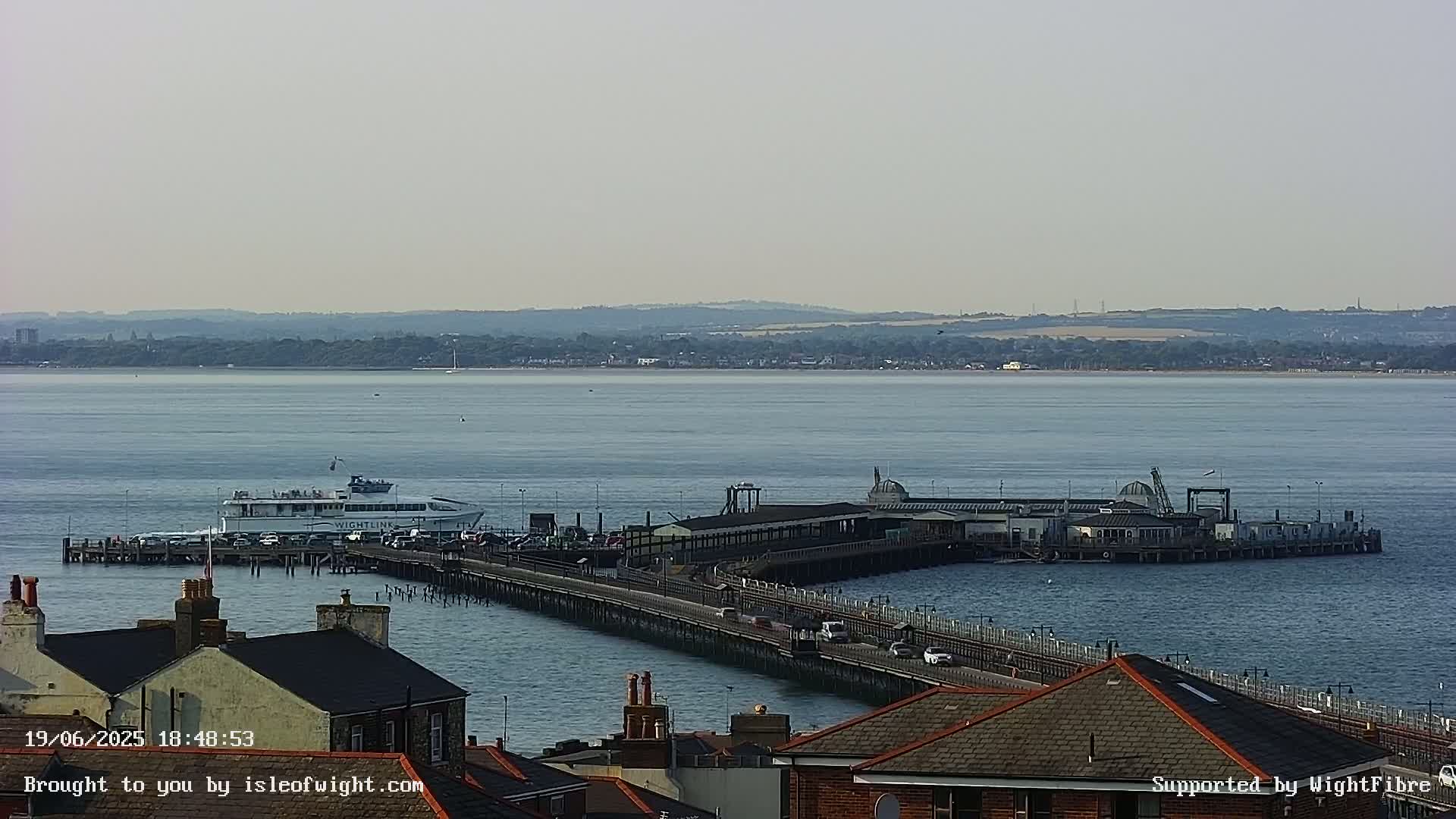 A ferry is docked at a long pier extending into a calm body of water, with a distant shoreline visible under a hazy sky.