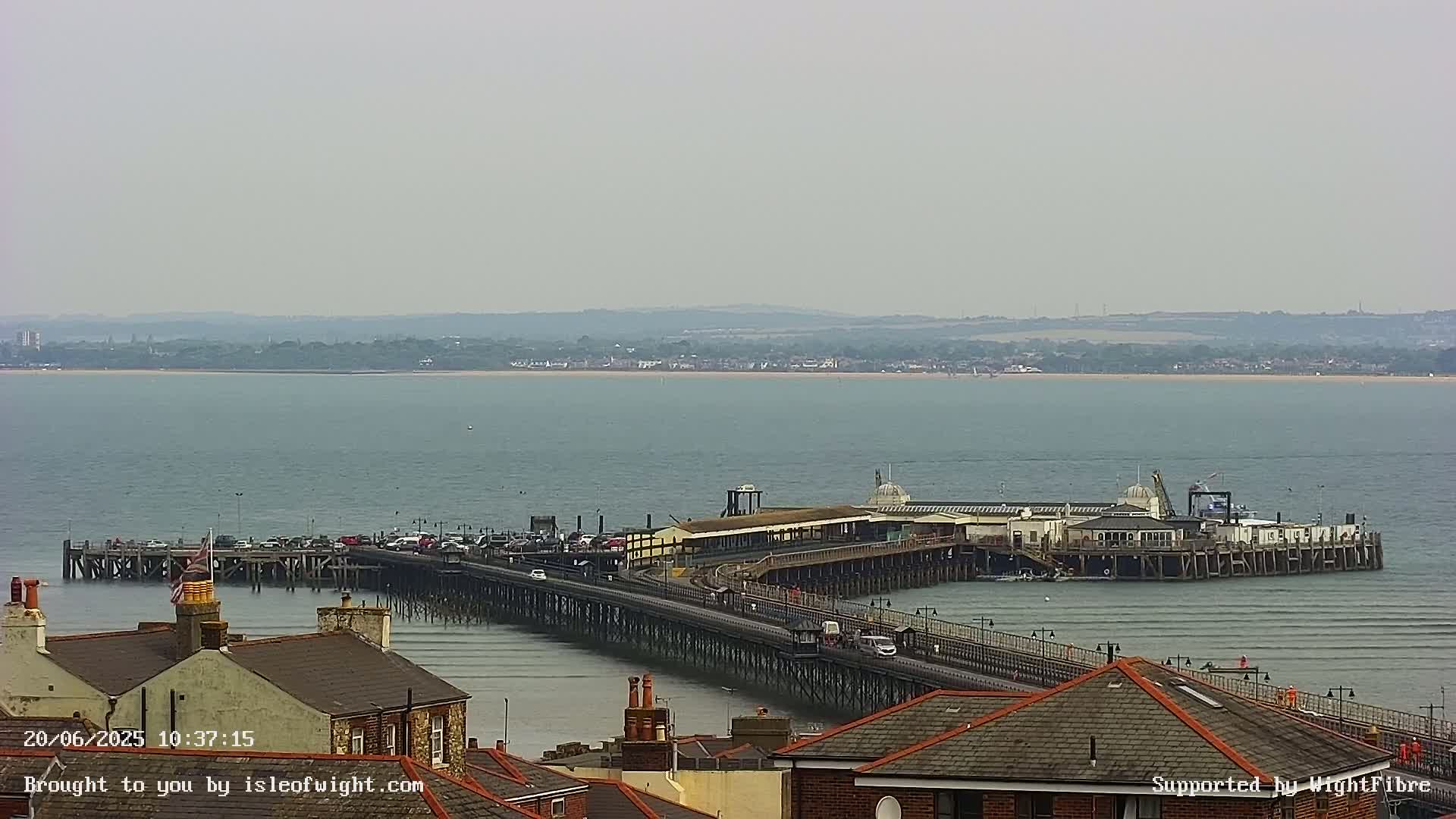 A long pier extends into a calm, gray-toned body of water, with buildings and rooftops visible in the foreground under an overcast sky.