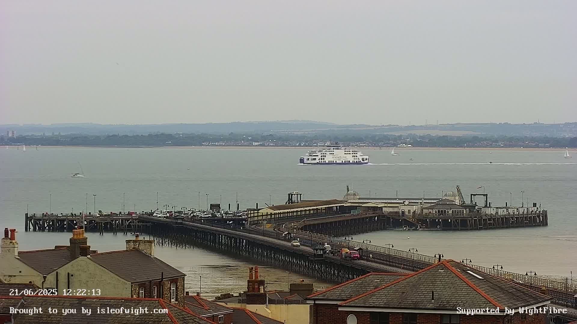 A ferry approaches a long pier extending into a calm, overcast body of water, with buildings and rooftops visible in the foreground.