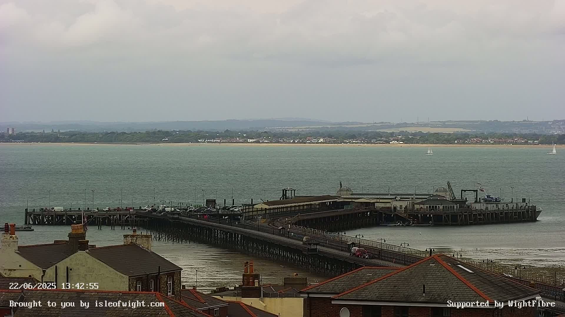 A long pier extends from a shoreline into a calm body of water under an overcast sky, with a distant coastline visible.