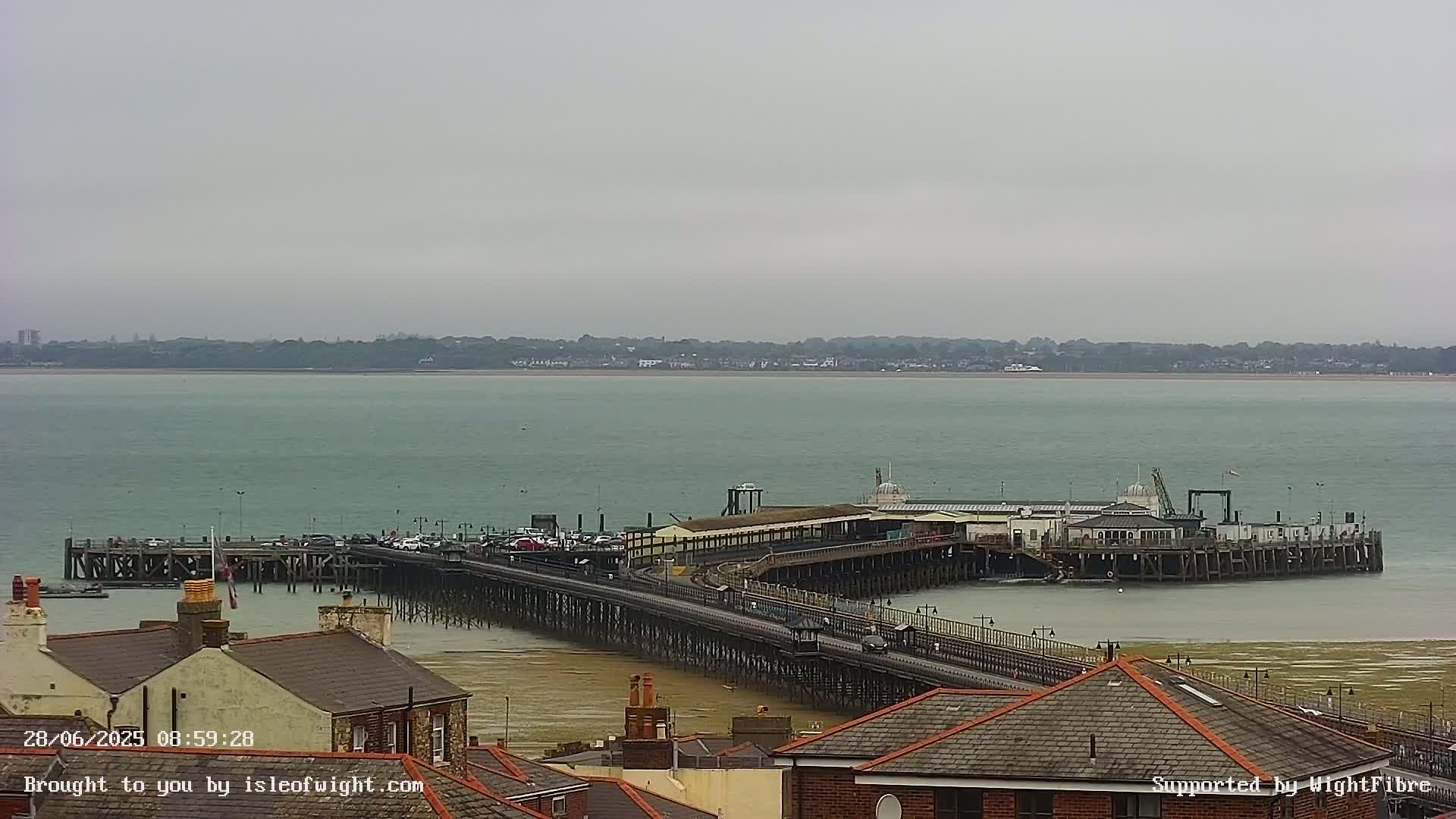 A long pier extends into a calm, gray-sky sea, with buildings visible in the foreground and a distant shoreline.
