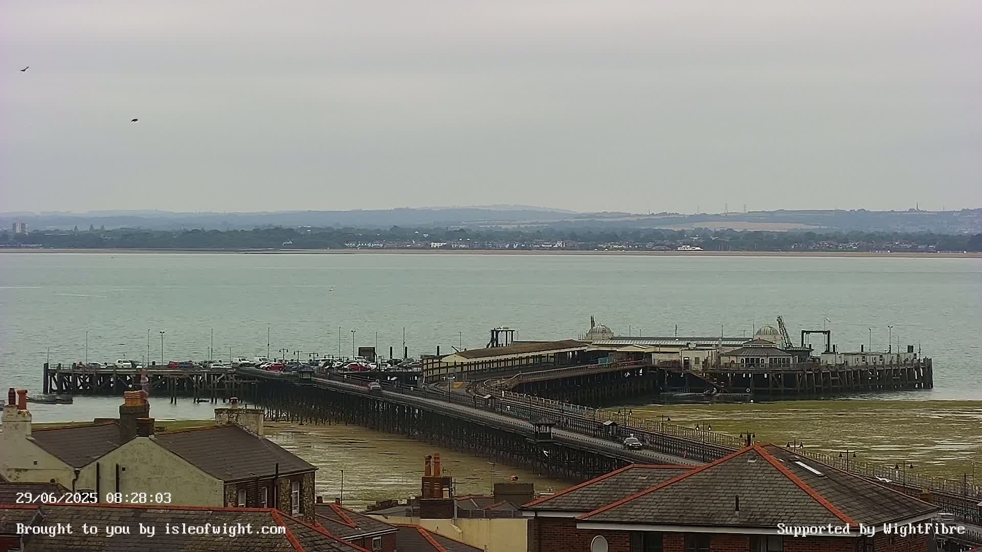 A long pier extends into a calm, grey sea under an overcast sky, with buildings and a distant shoreline visible.