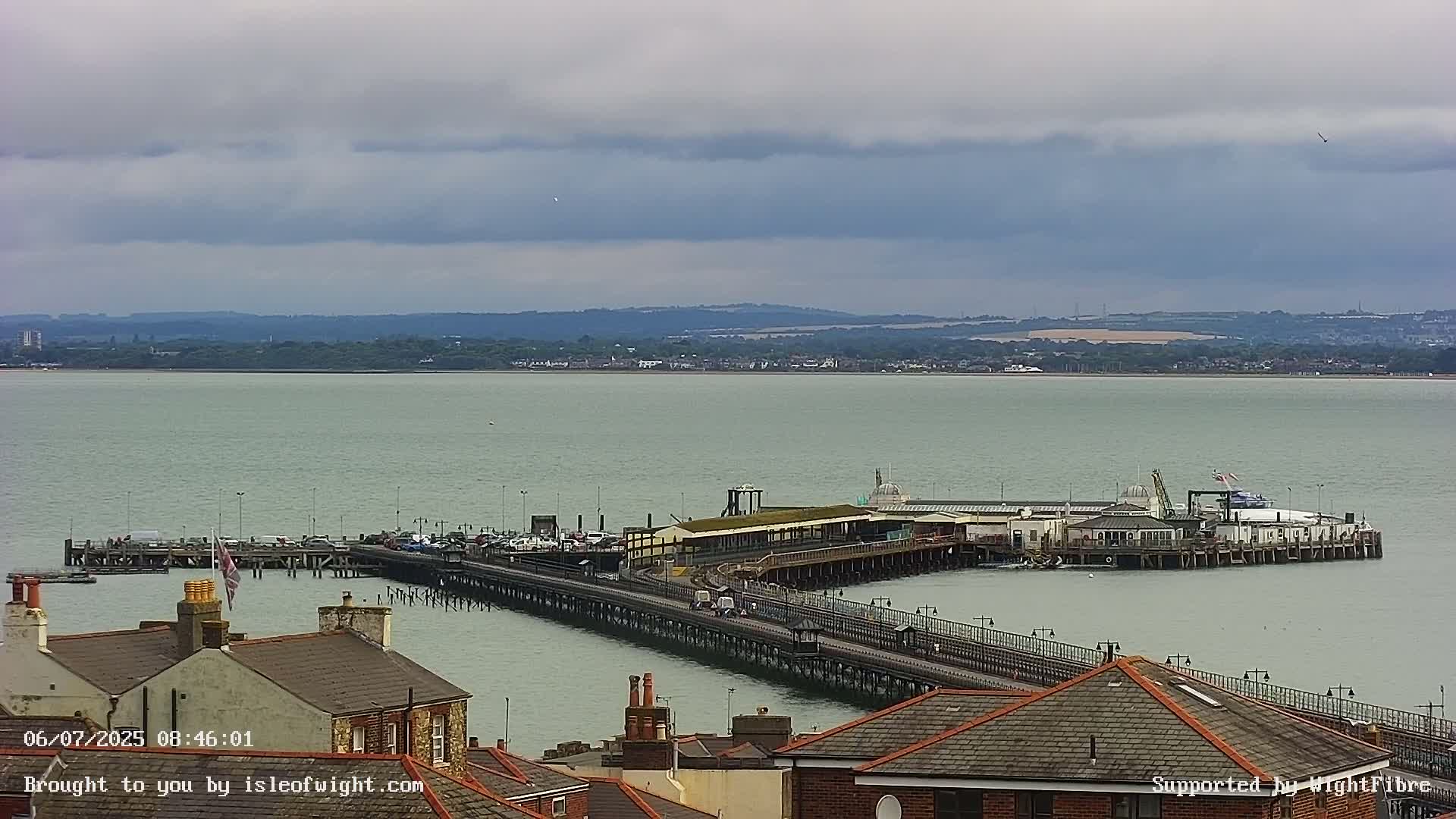 A long pier extends from a shoreline into calm, gray water under an overcast sky, with buildings and a distant coastline visible.