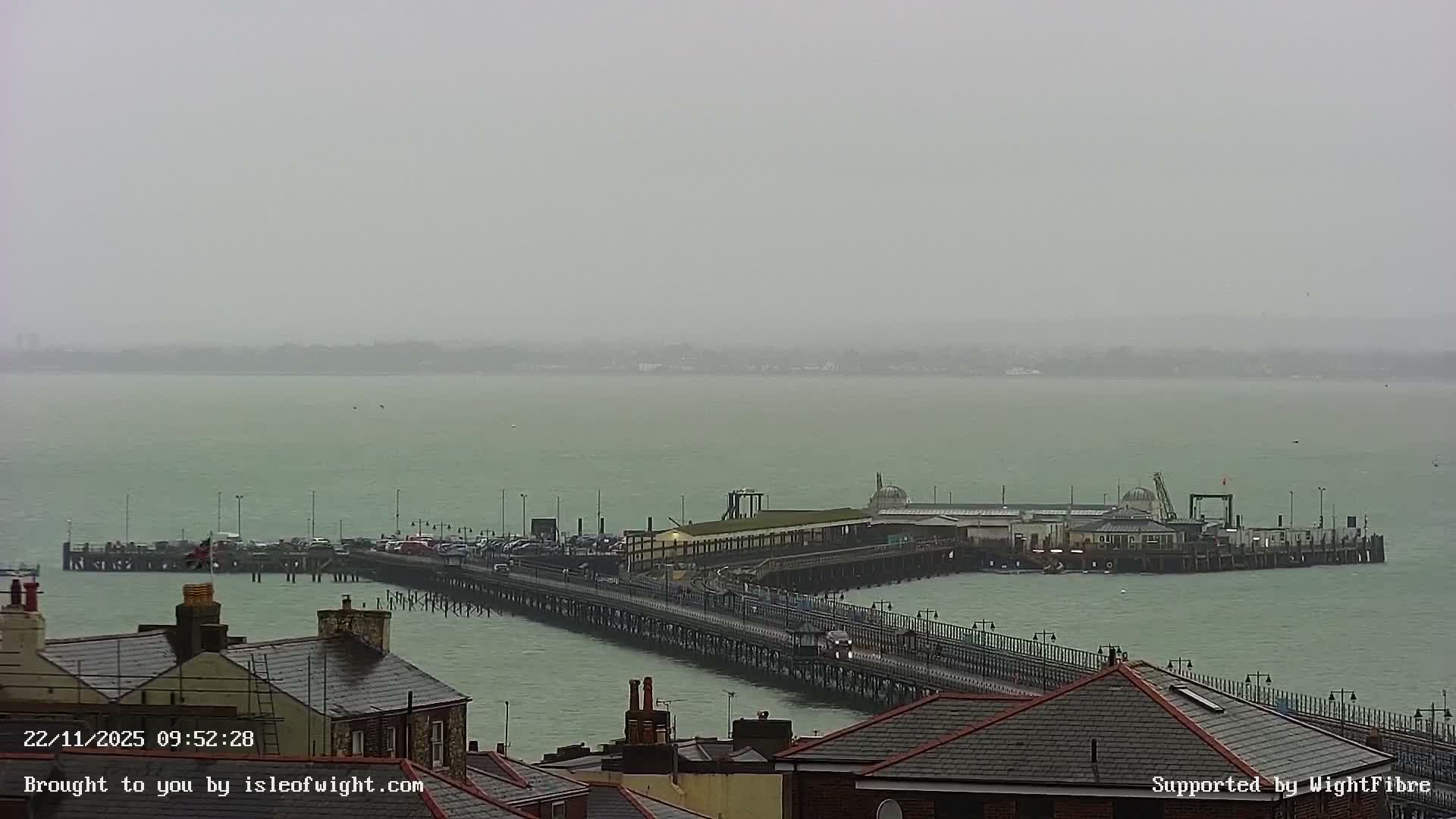 An elevated view shows a long pier, featuring buildings, parked cars, and train tracks, extending into a green-tinged body of water towards a faint, hazy horizon, all beneath a uniform overcast sky.