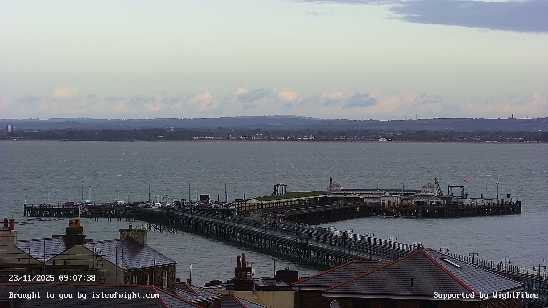 A long pier with buildings and vehicles extends into a choppy bay or sea, framed by rooftops in the foreground and a distant coastline with towns and hills under a partly cloudy sky.
