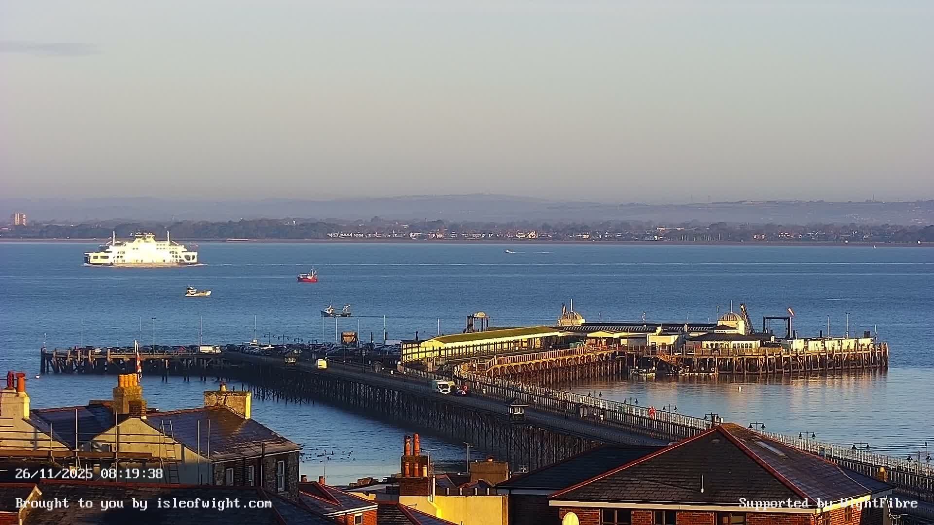 A clear, calm day reveals a wide body of water with a large white ferry and several smaller boats, framed by a long pier bustling with vehicles and buildings in the mid-ground, and residential roofs in the foreground, all set against a hazy distant shoreline and hills.