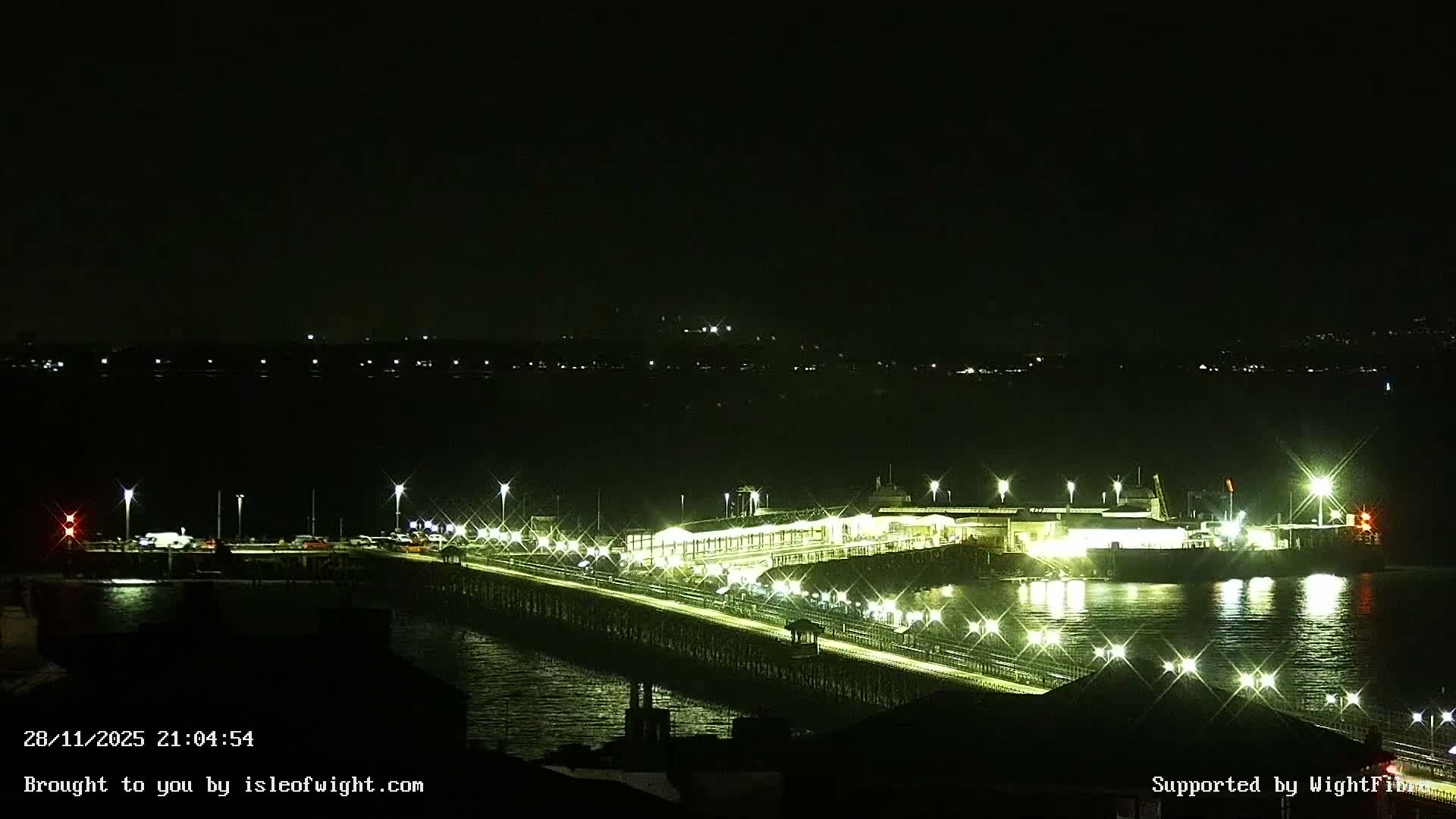 The image captures a clear, dark night over a brightly lit ferry terminal and pier, with a ferry docked and distant lights visible across the water.