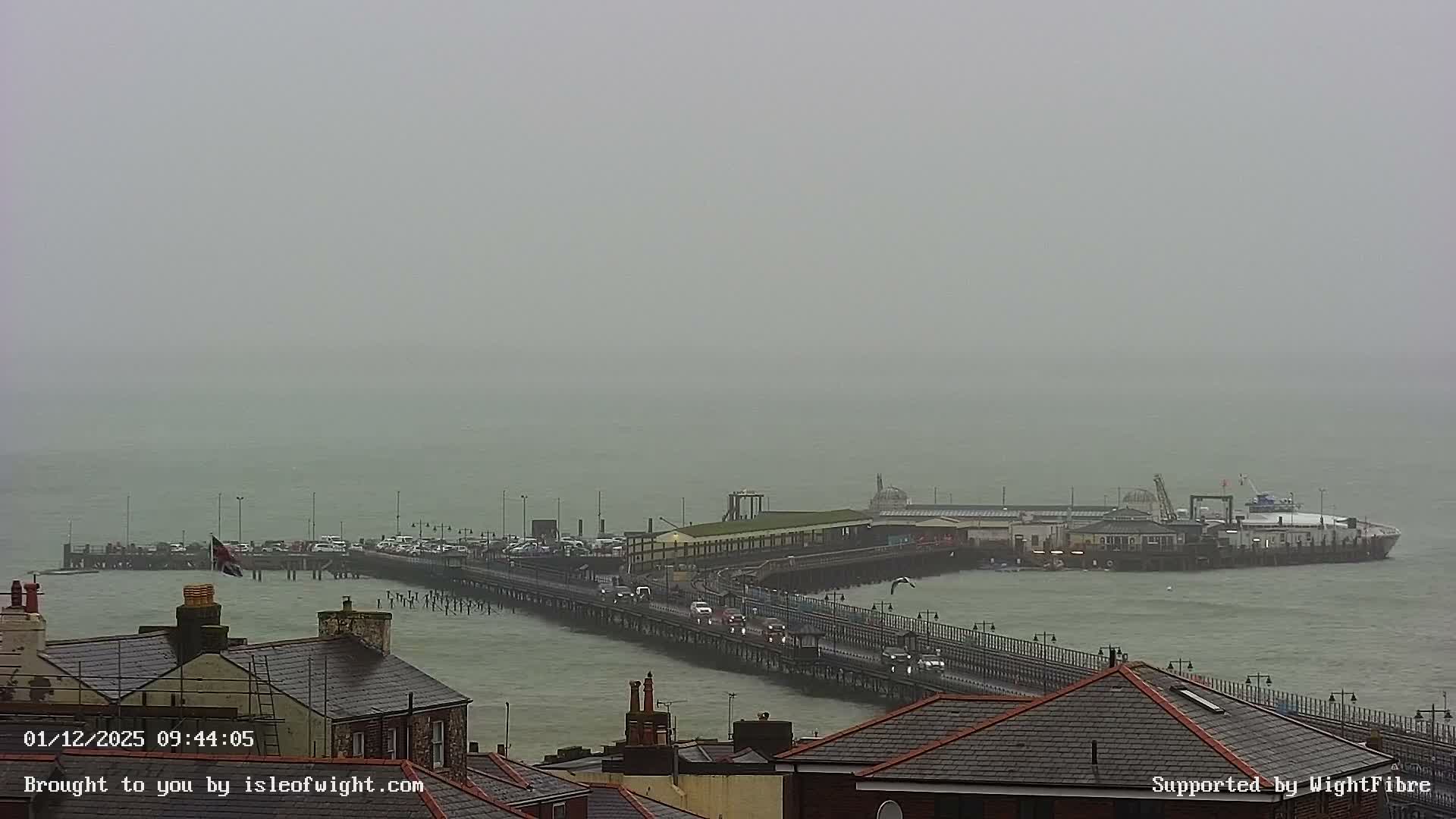 A long pier with cars and waterfront buildings extends into a choppy grey sea, where a large ferry is docked at its end, all under a heavily overcast and hazy sky.