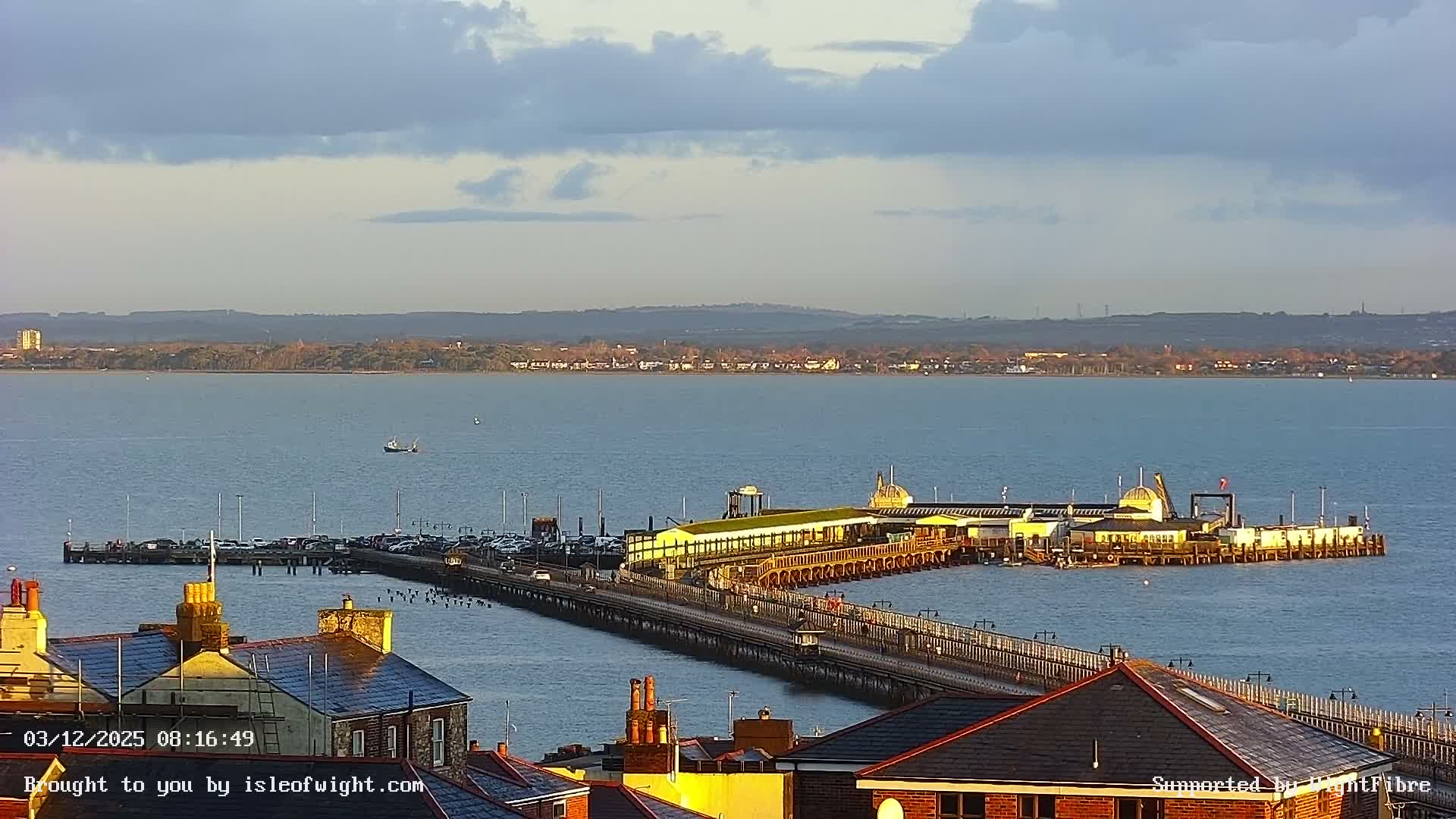 A long pier with buildings and a busy car park extends into calm blue waters under a partly cloudy morning sky, with distant land visible across the bay and residential rooftops in the immediate foreground bathed in warm sunlight.