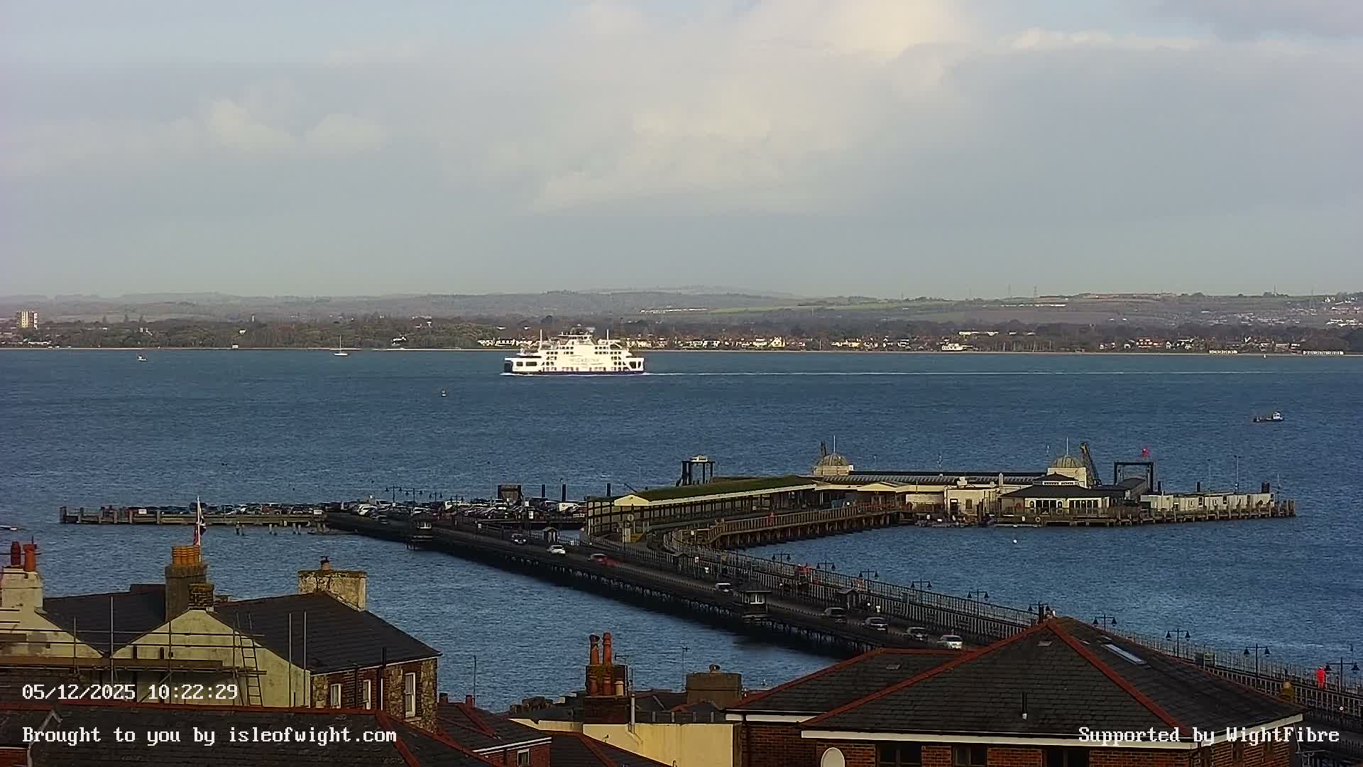 A large white ferry traverses a calm blue bay under a partly cloudy sky, with a long pier extending from the foreground into the water and a distant tree-lined coastline.