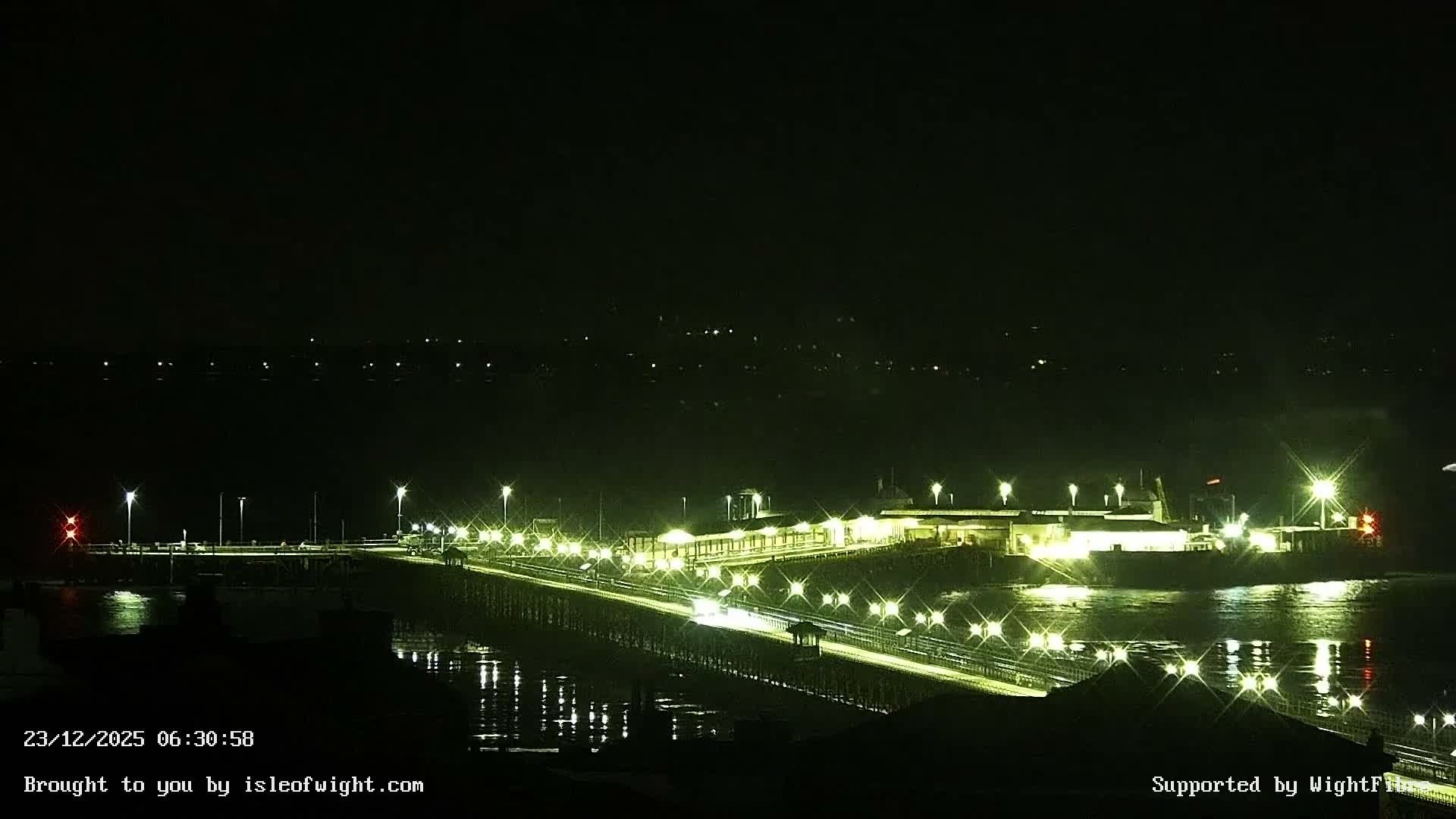 A brightly lit pier or causeway extends across calm dark water towards illuminated buildings and a ferry terminal at night, under a clear sky with distant lights visible on the horizon.