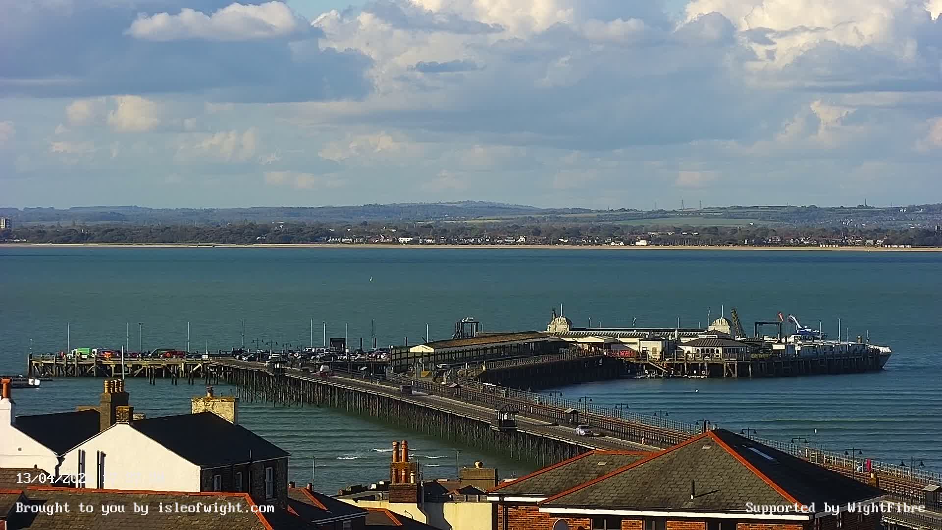 A brightly lit pier or causeway extends across calm dark water towards illuminated buildings and a ferry terminal at night, under a clear sky with distant lights visible on the horizon.
