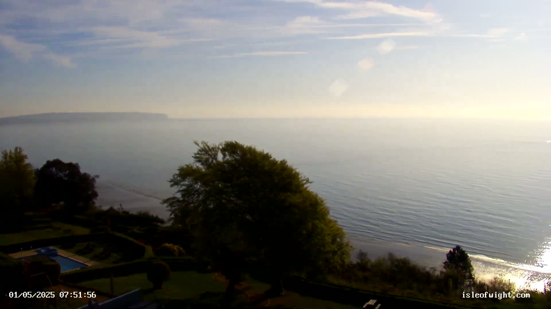 A hazy, calm sea stretches to a distant, indistinct coastline under a pale blue sky, viewed from a vantage point overlooking a manicured lawn and swimming pool.