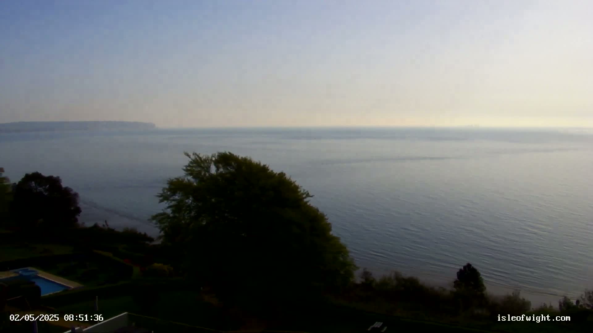 A hazy, calm ocean is seen from a high vantage point, with green trees and a swimming pool below, and land visible in the distance.
