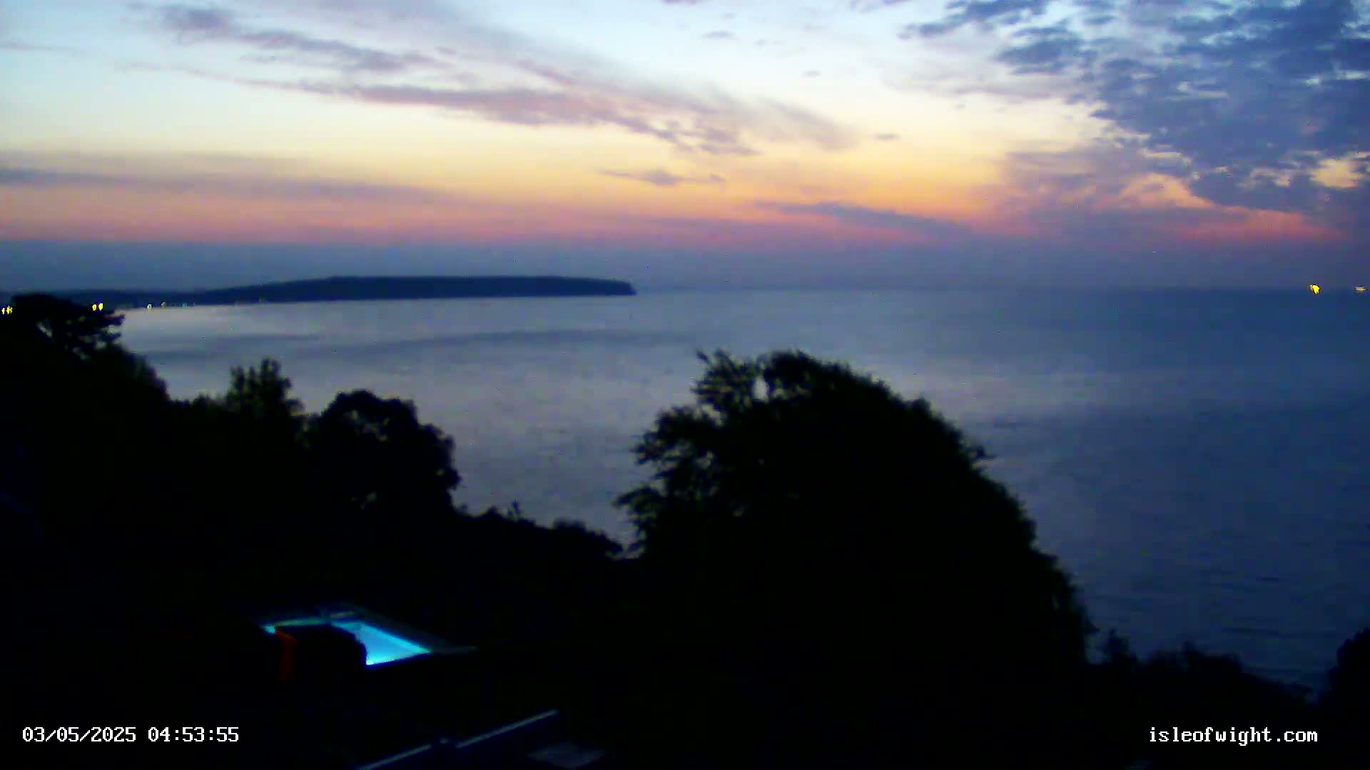 A calm body of water stretches to a distant landmass under a twilight sky, with dark silhouettes of trees and a pool visible in the foreground.