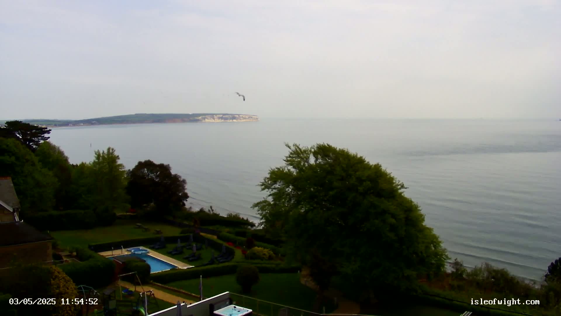 A tranquil ocean scene with a distant island is viewed from a high vantage point overlooking a manicured lawn containing a swimming pool and hot tub, under an overcast sky.