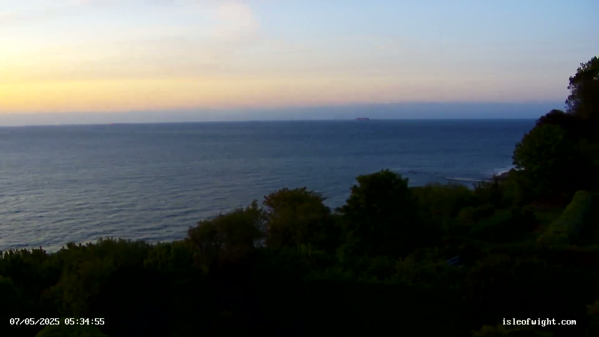A tranquil ocean scene at sunset, viewed from a high vantage point overlooking dark green vegetation.