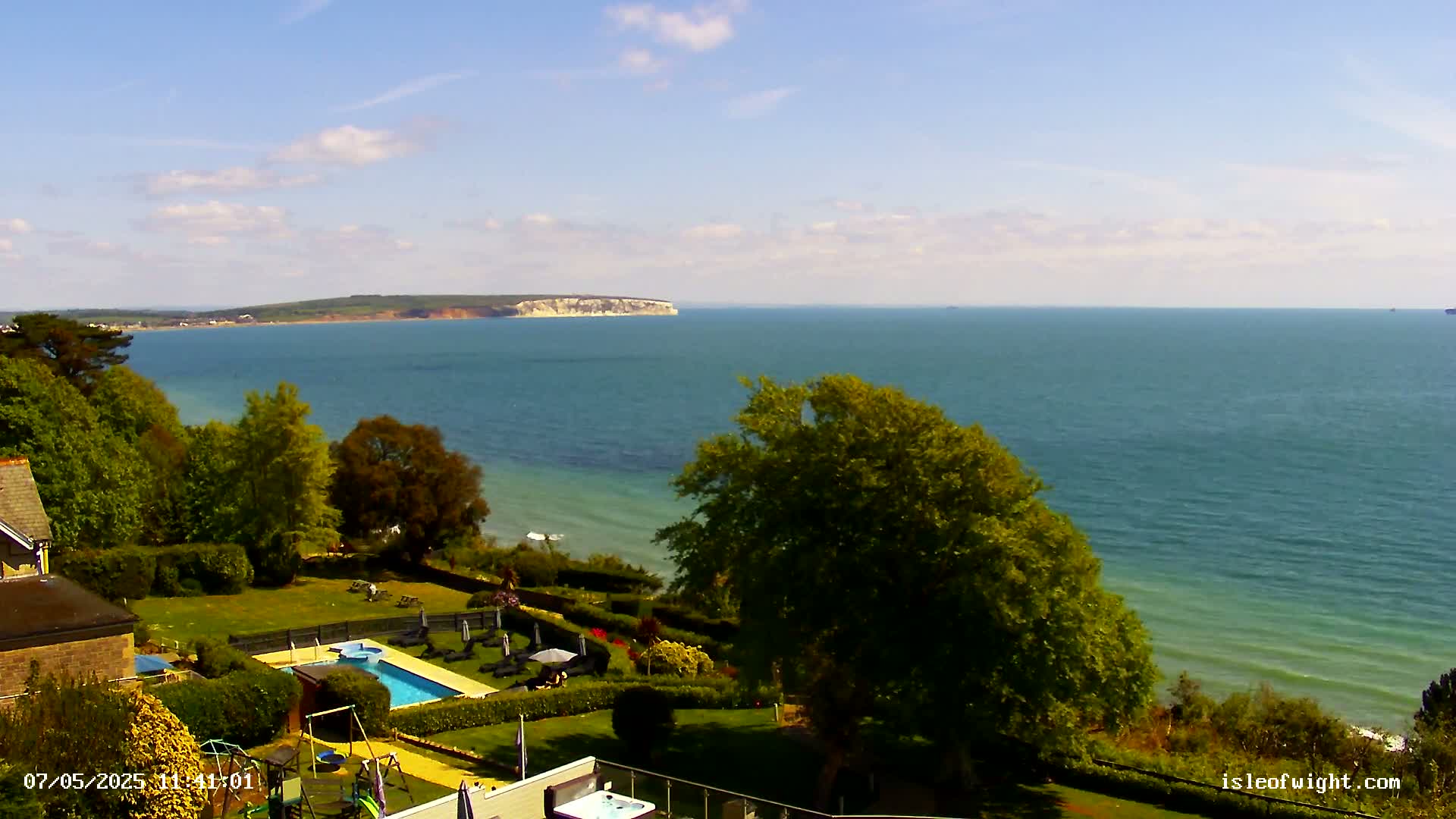 A swimming pool and playground are situated on a grassy hill overlooking a calm ocean and a distant landmass under a mostly sunny sky.