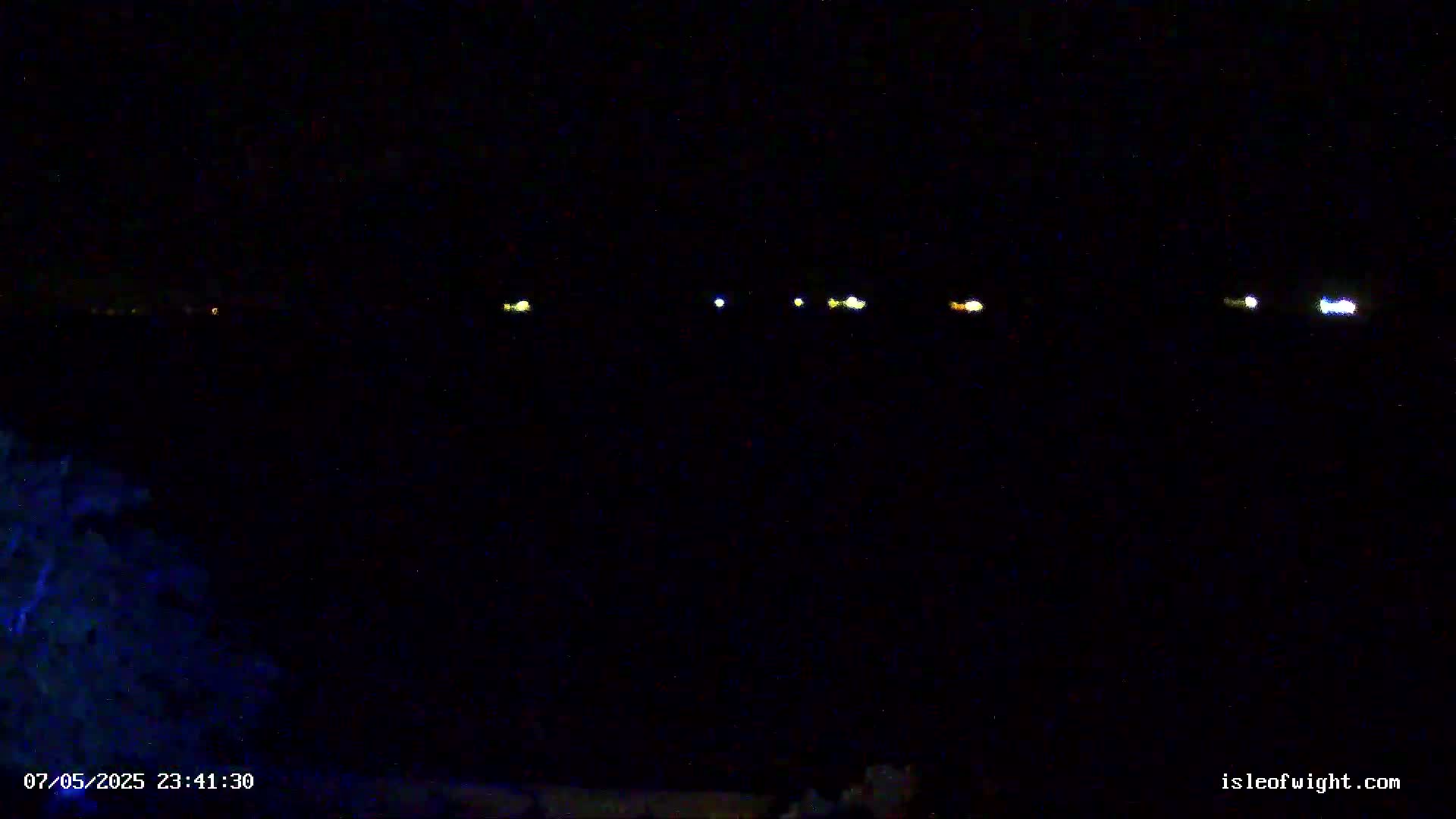 A dark nighttime scene shows a line of distant lights across a black body of water, with dark vegetation visible in the bottom left corner.