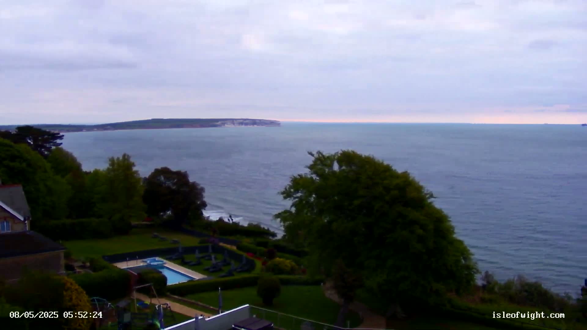 A coastal scene under an overcast sky shows a swimming pool and lawn next to a building overlooking a calm sea and distant land.