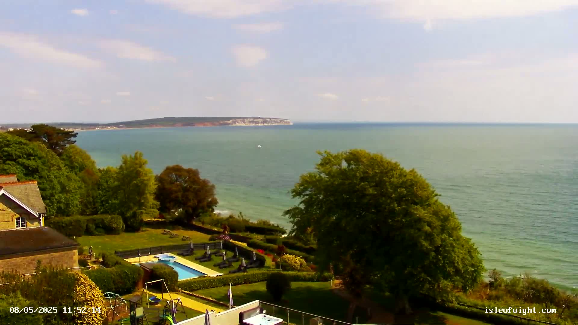 A seaside property with a swimming pool and playground overlooks a calm ocean and distant land under a partly cloudy sky.
