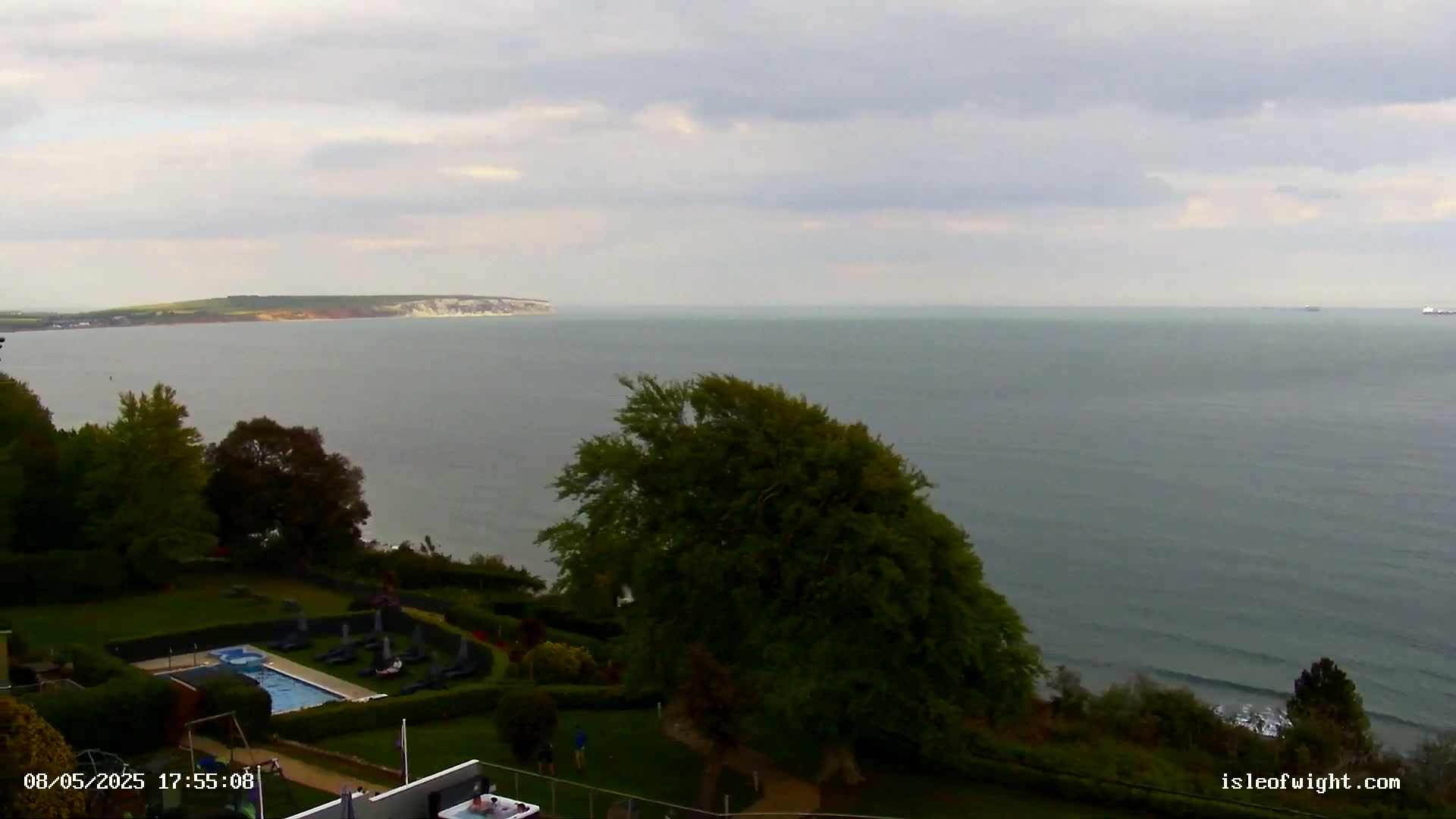 An overcast day reveals a coastal view of a calm sea, a swimming pool nestled in a grassy area with landscaping, and a distant landmass.