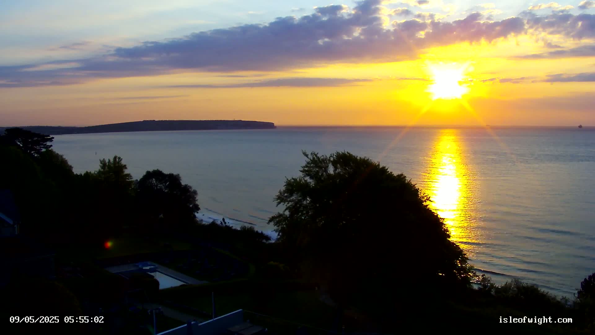 A vibrant sunrise casts a golden glow on the calm ocean, illuminating a distant headland and silhouetting trees on a coastal hillside.