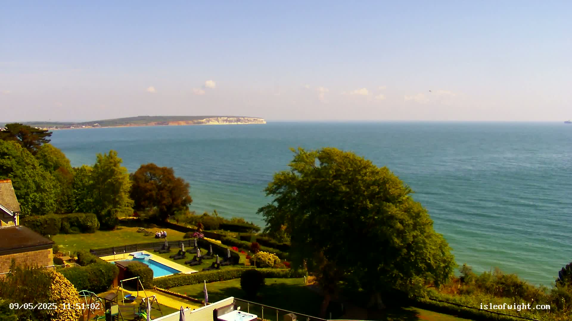 A sunny coastal scene shows a swimming pool and lawn next to a building overlooking a calm sea and distant cliffs.