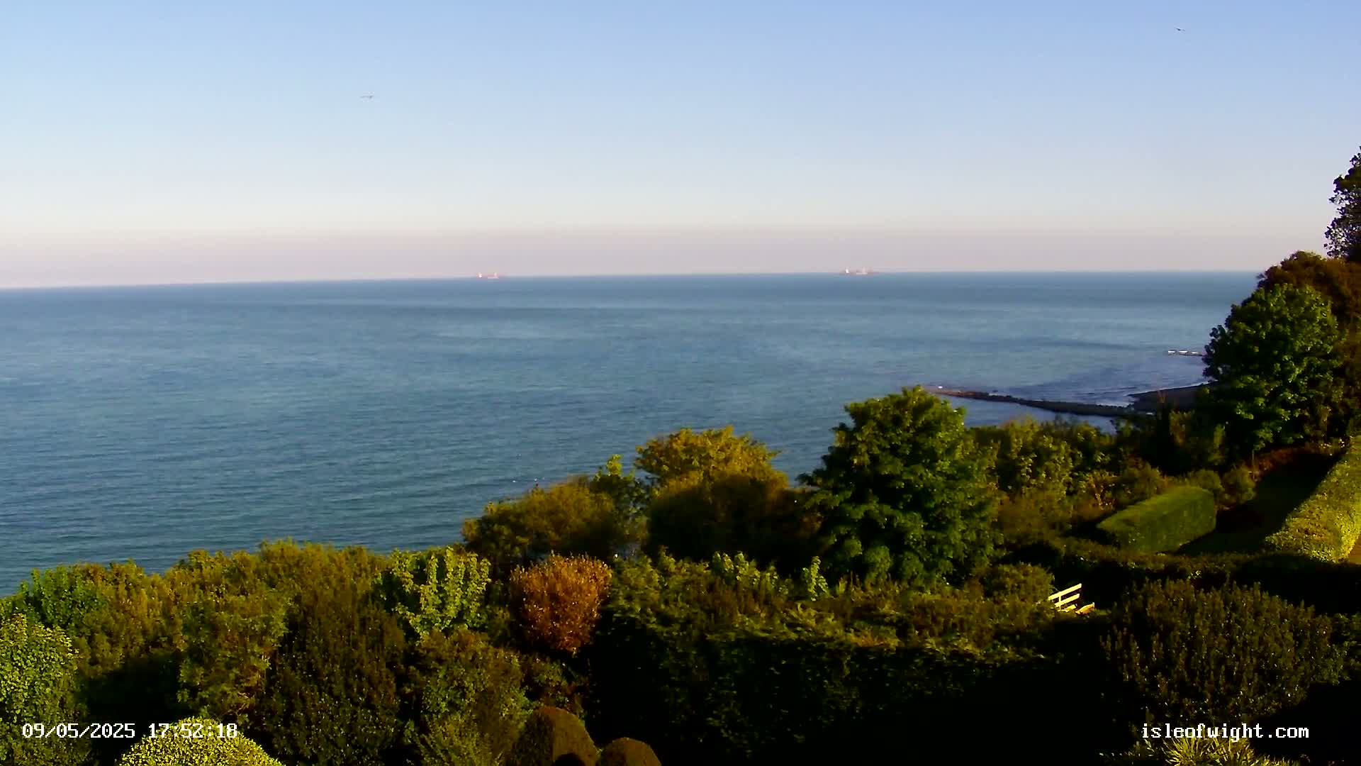 A calm, blue ocean stretches to the horizon under a clear sky, viewed from a high vantage point overlooking a lush green landscape of trees and shrubs.