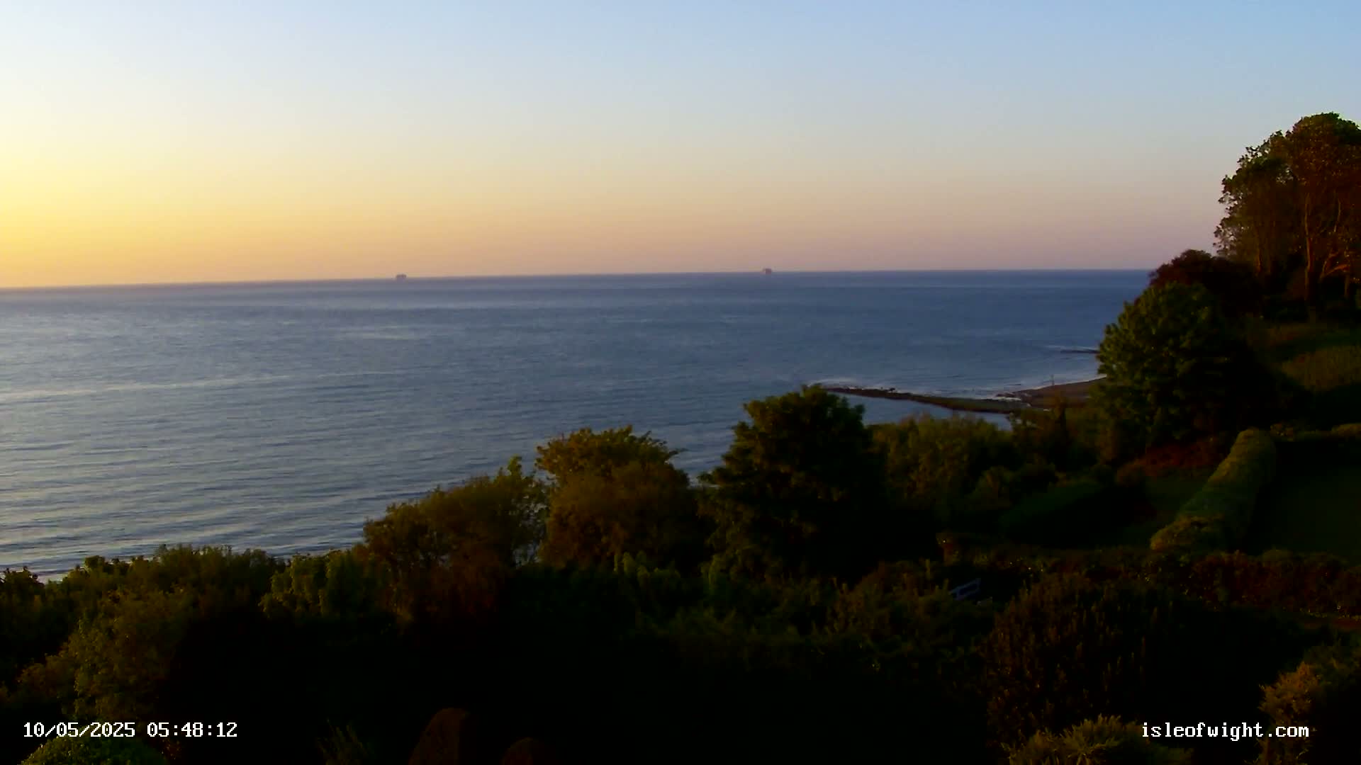 A calm ocean at sunset is viewed from a high vantage point overlooking a lush, green hillside.