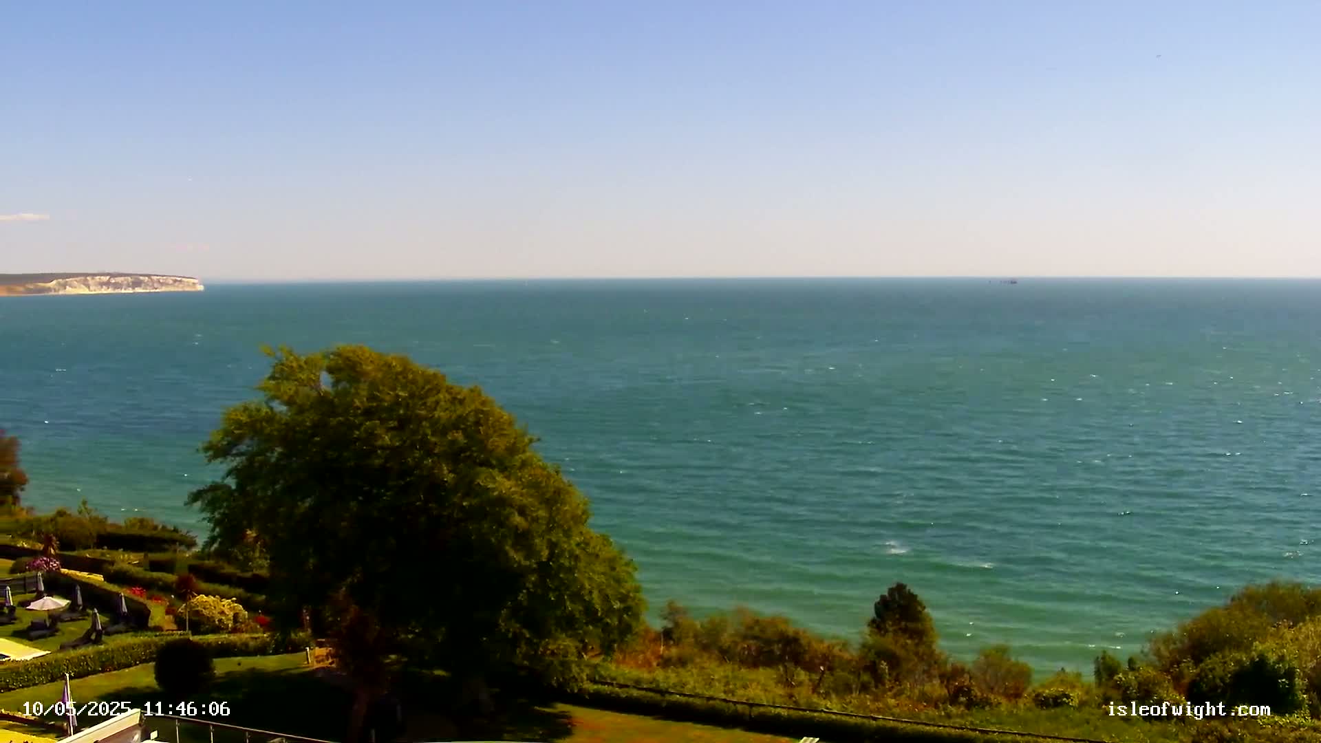 A tranquil ocean scene on a sunny day shows a large body of water stretching to the horizon, with a distant cliff face and lush green vegetation in the foreground.