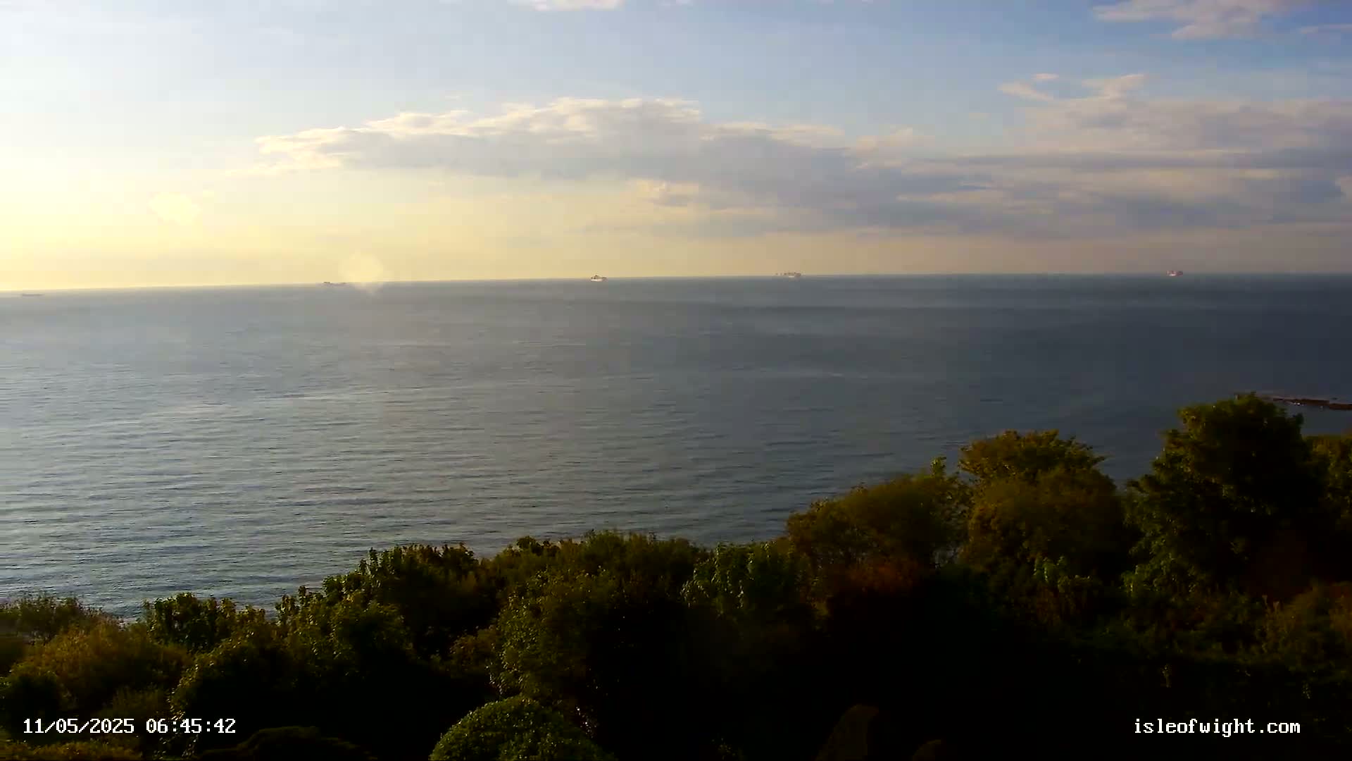 A calm sea under a partly cloudy sky is viewed from a high vantage point overlooking a line of dark green vegetation.