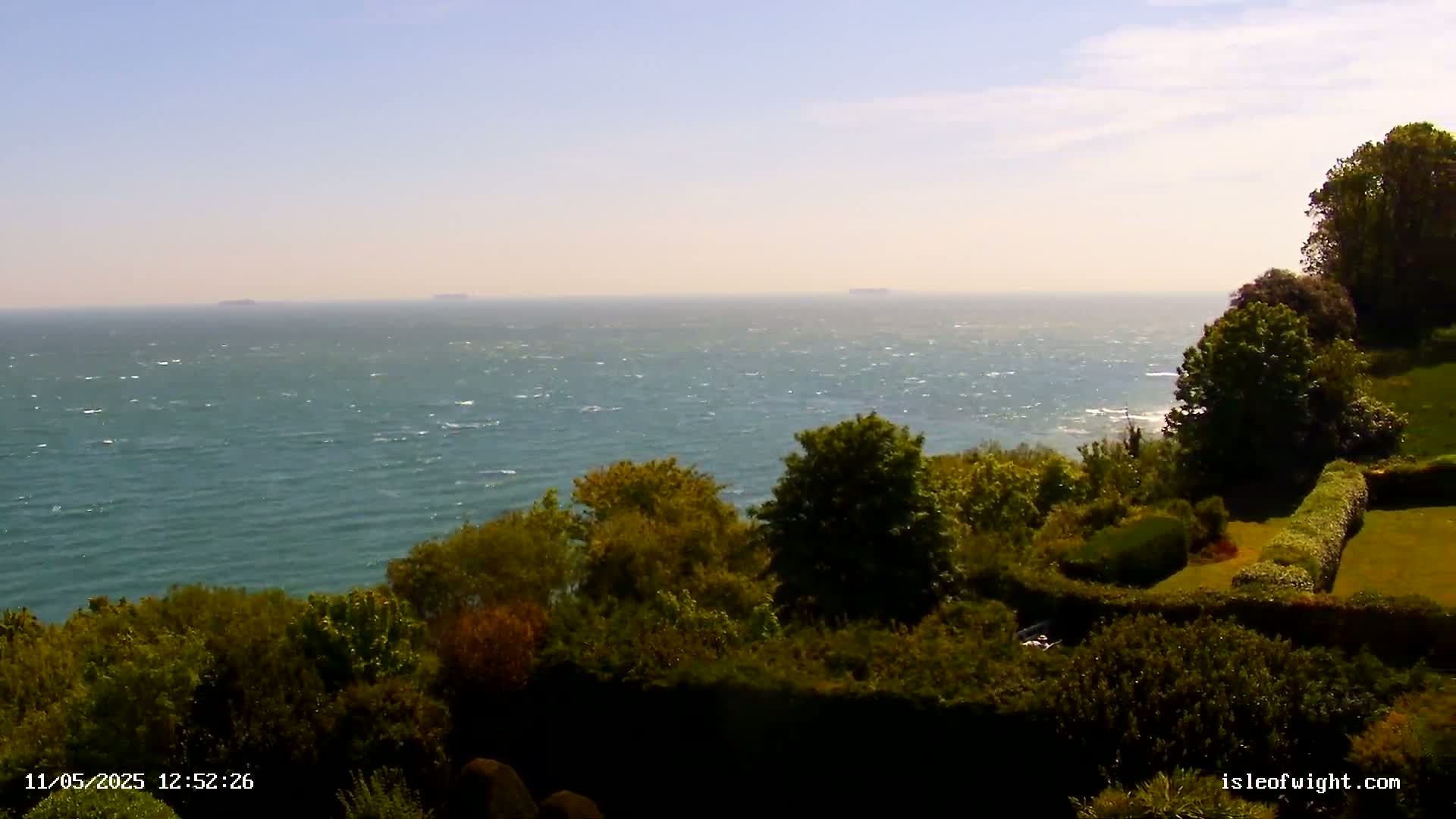 A windy, sunny day reveals a vast expanse of blue ocean dotted with distant ships, viewed from a verdant clifftop with lush green foliage and manicured hedges.