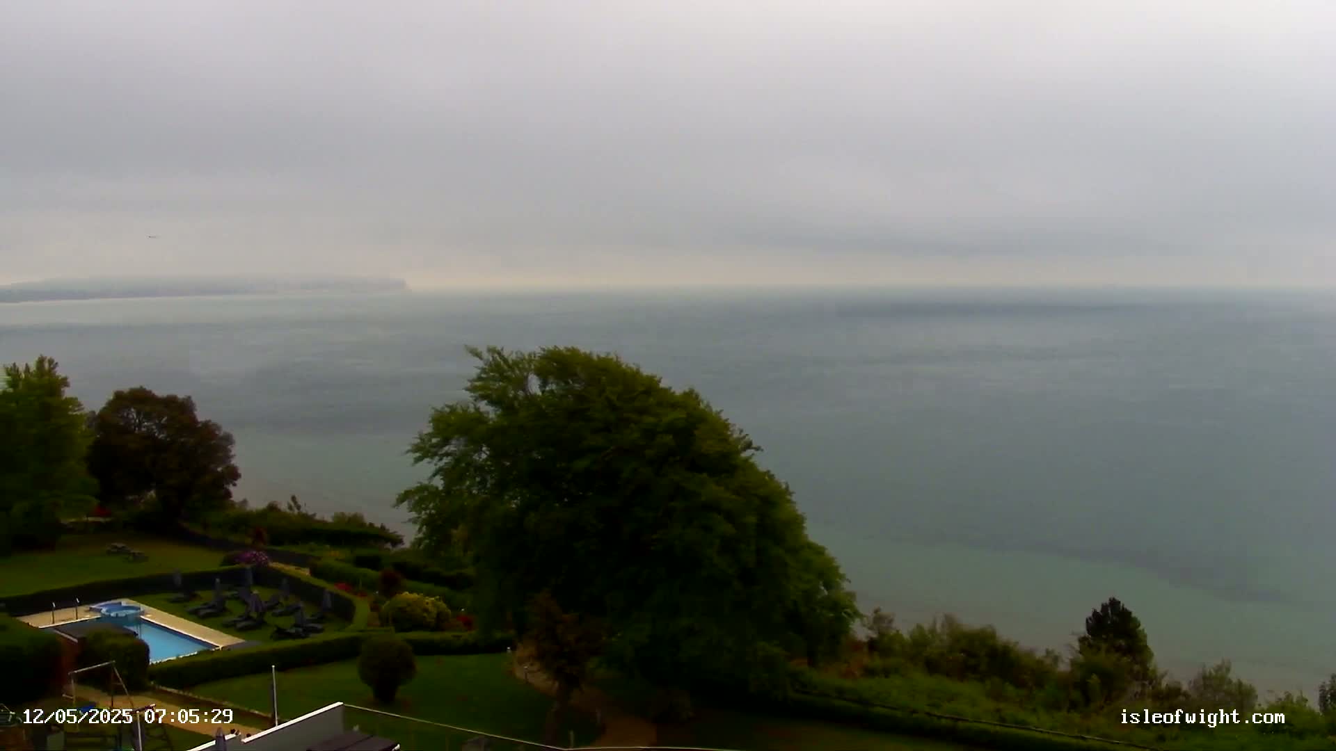 A hazy, overcast day reveals a calm body of water stretching to a distant shoreline, viewed from an elevated vantage point featuring a swimming pool and manicured lawns.