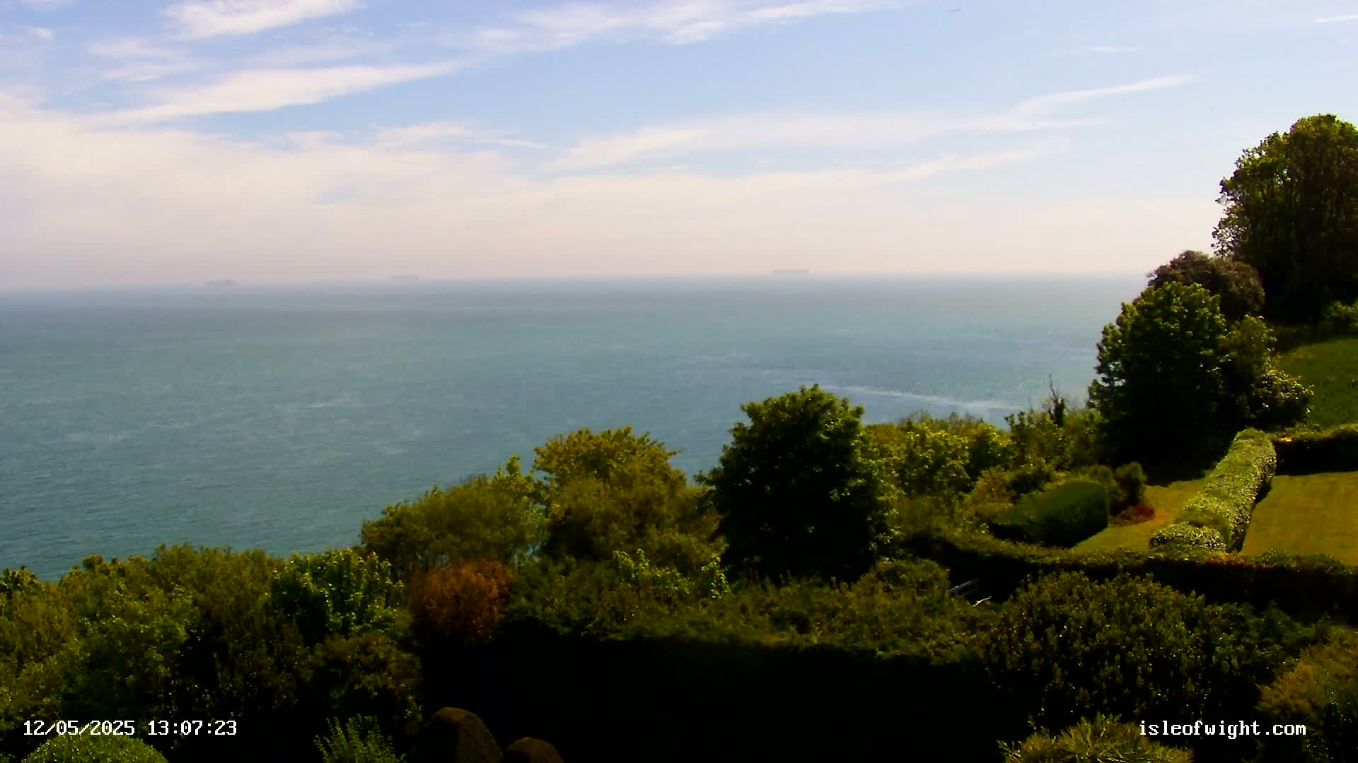 A vast expanse of calm, teal-colored ocean stretches to the horizon under a mostly clear, light-blue sky, viewed from a verdant clifftop dotted with lush green trees and shrubs.