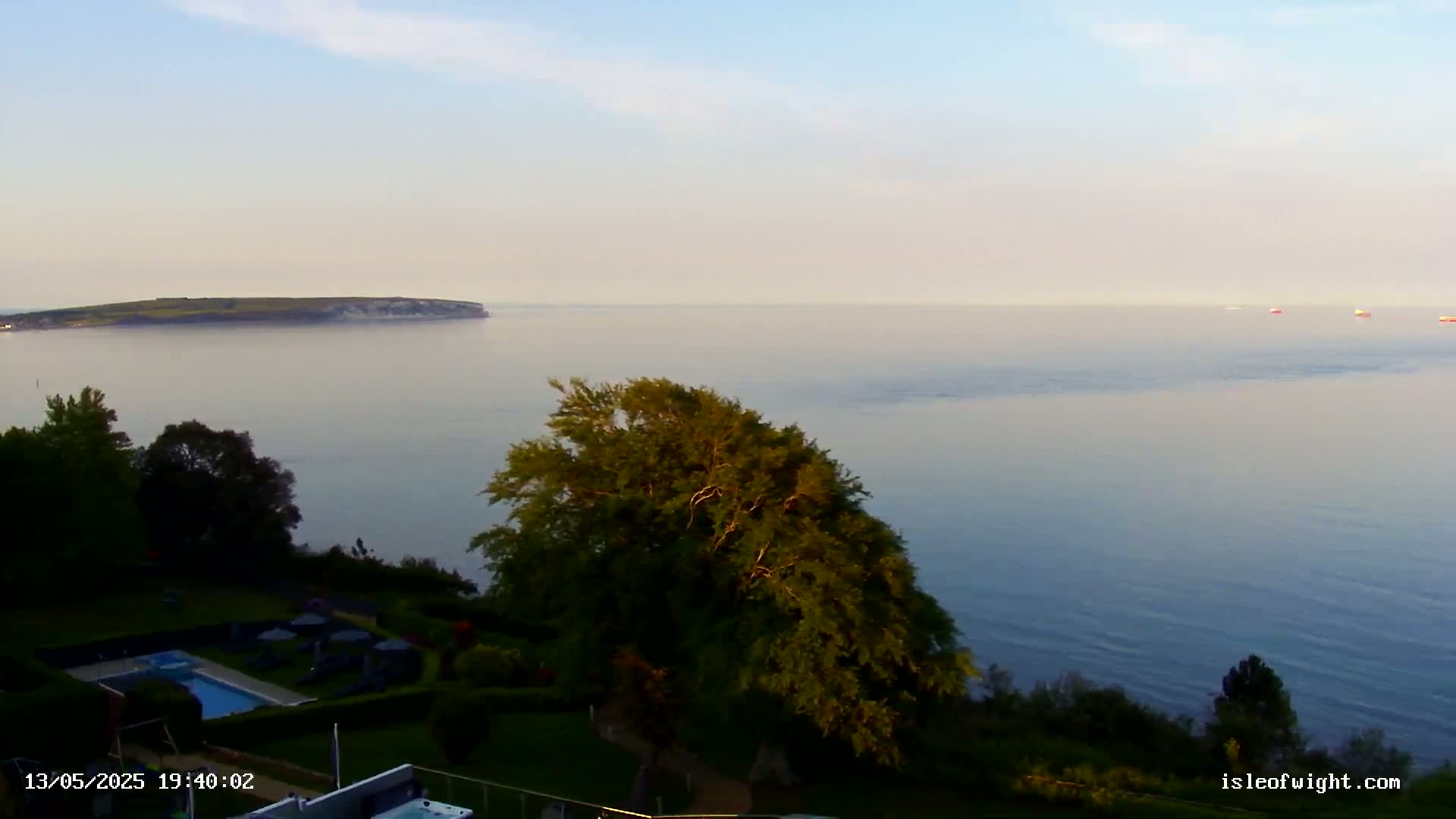 A calm, hazy evening overlooks a body of water with a distant island and several ships, showcasing a swimming pool and lush greenery in the foreground.