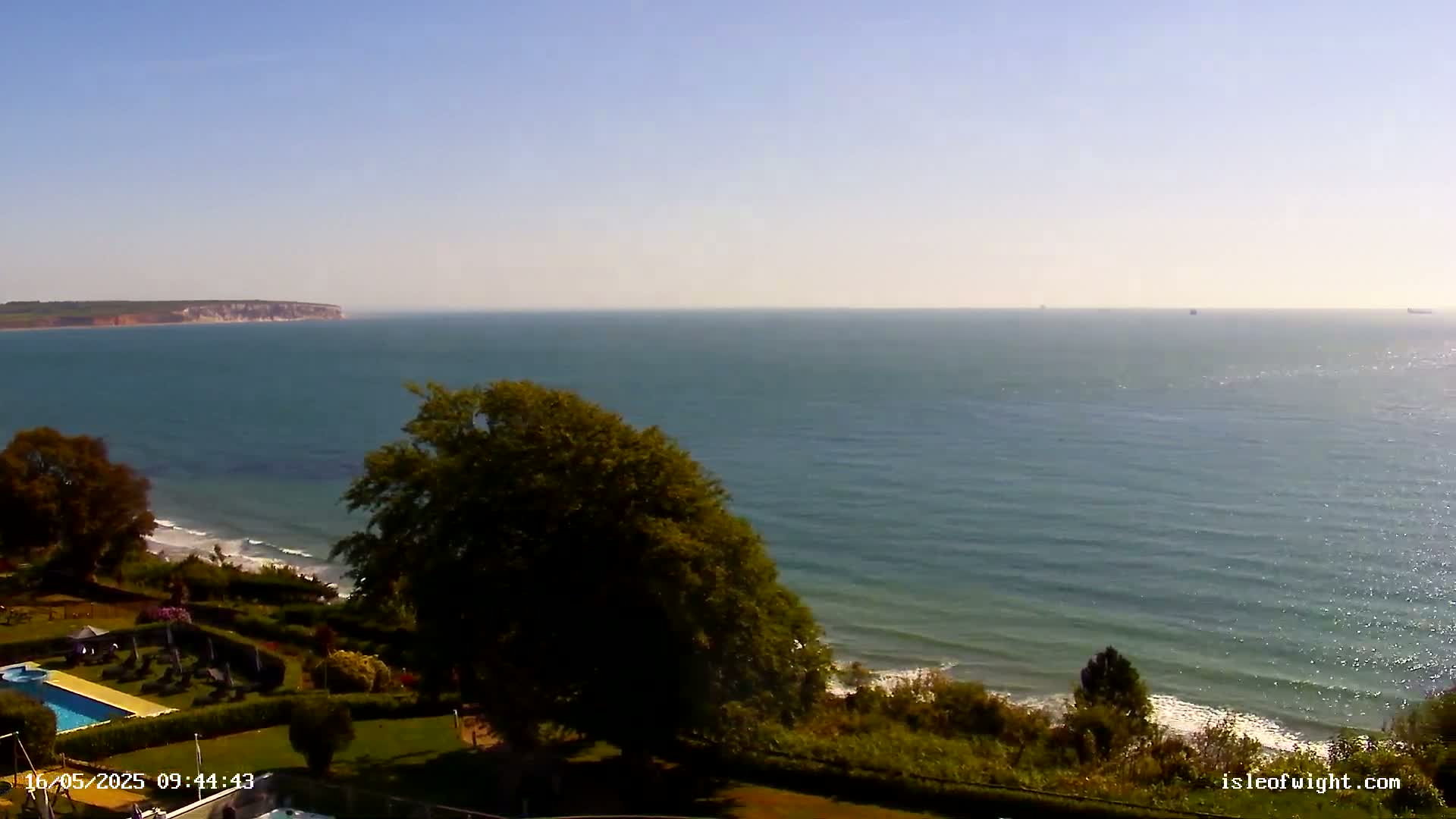 A tranquil ocean scene on a sunny day shows a calm sea with gentle waves lapping a shoreline bordered by lush green vegetation and a swimming pool.