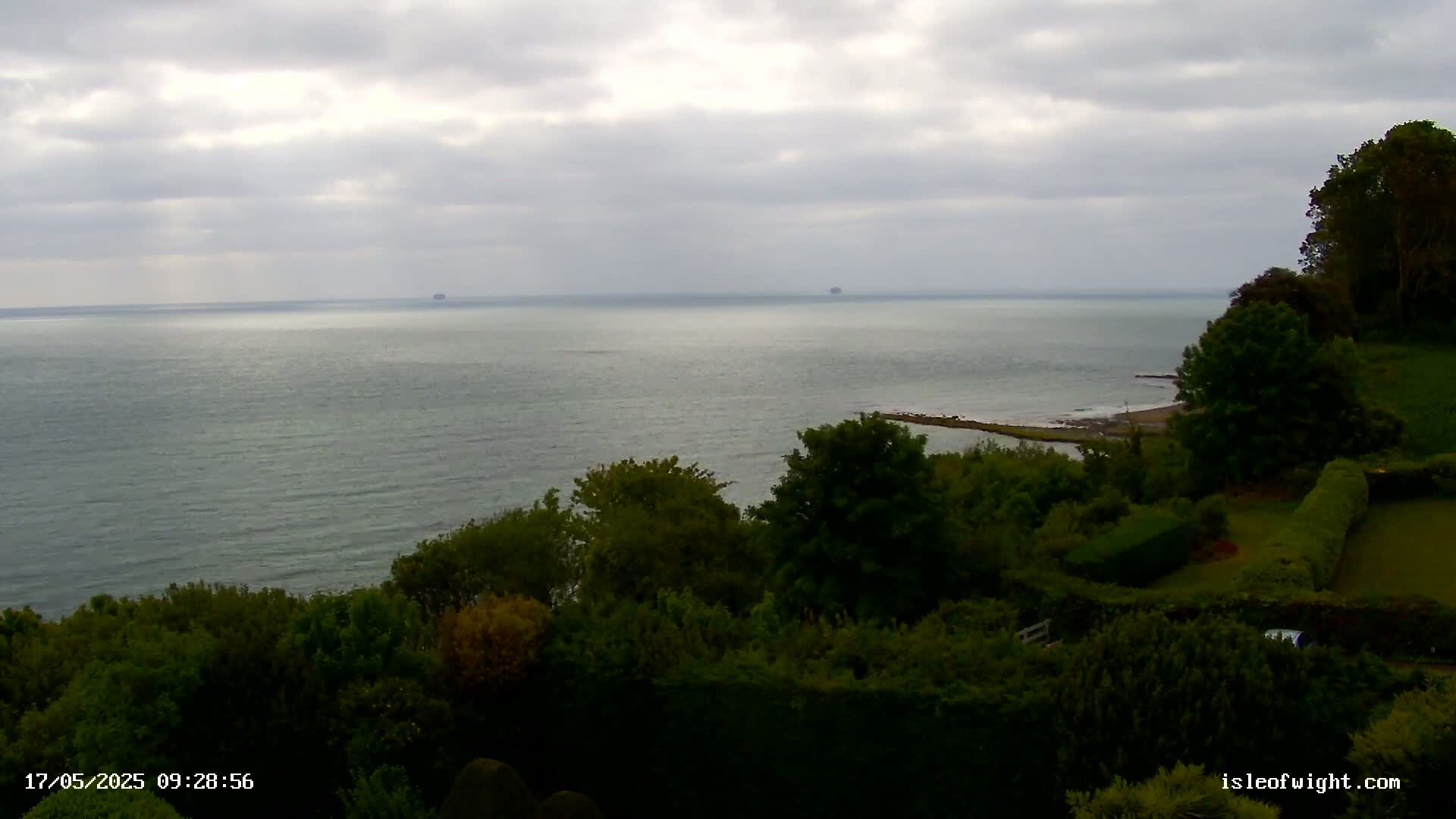 A calm, gray-sky day overlooks a tranquil sea from a verdant clifftop dotted with lush green trees and shrubbery.