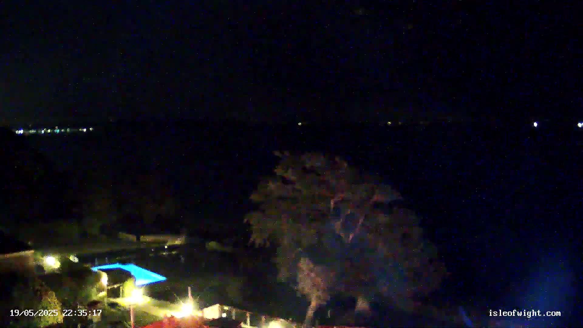 A nighttime view shows a dark body of water with faint lights on the horizon, a tree silhouetted against the darkness, and a pool illuminated by blue lights in the foreground.