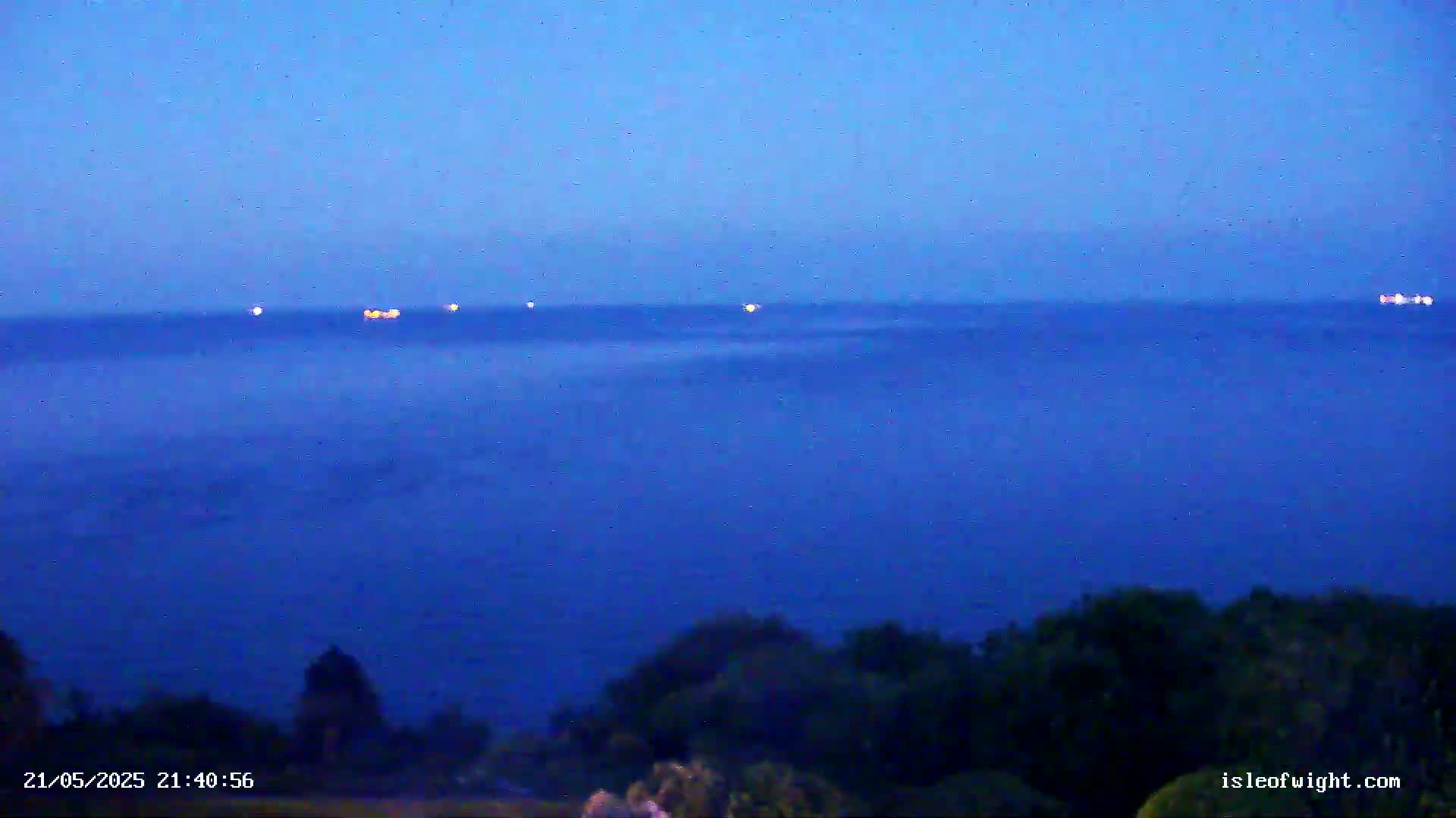 Several ships with illuminated lights are visible on a dark blue, seemingly calm sea at twilight, with dark green land in the foreground.