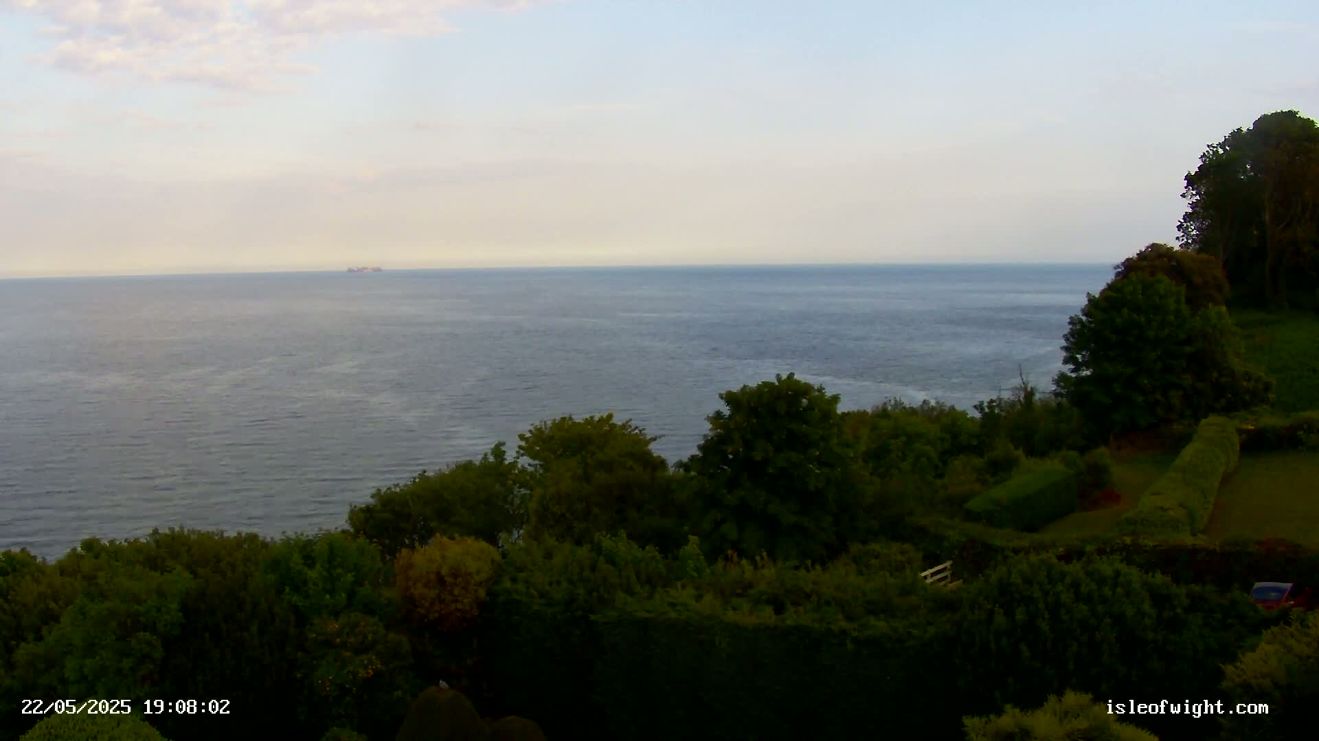 A calm sea stretches to the horizon under a pale sky, viewed from a verdant clifftop overlooking lush greenery and neatly trimmed hedges.